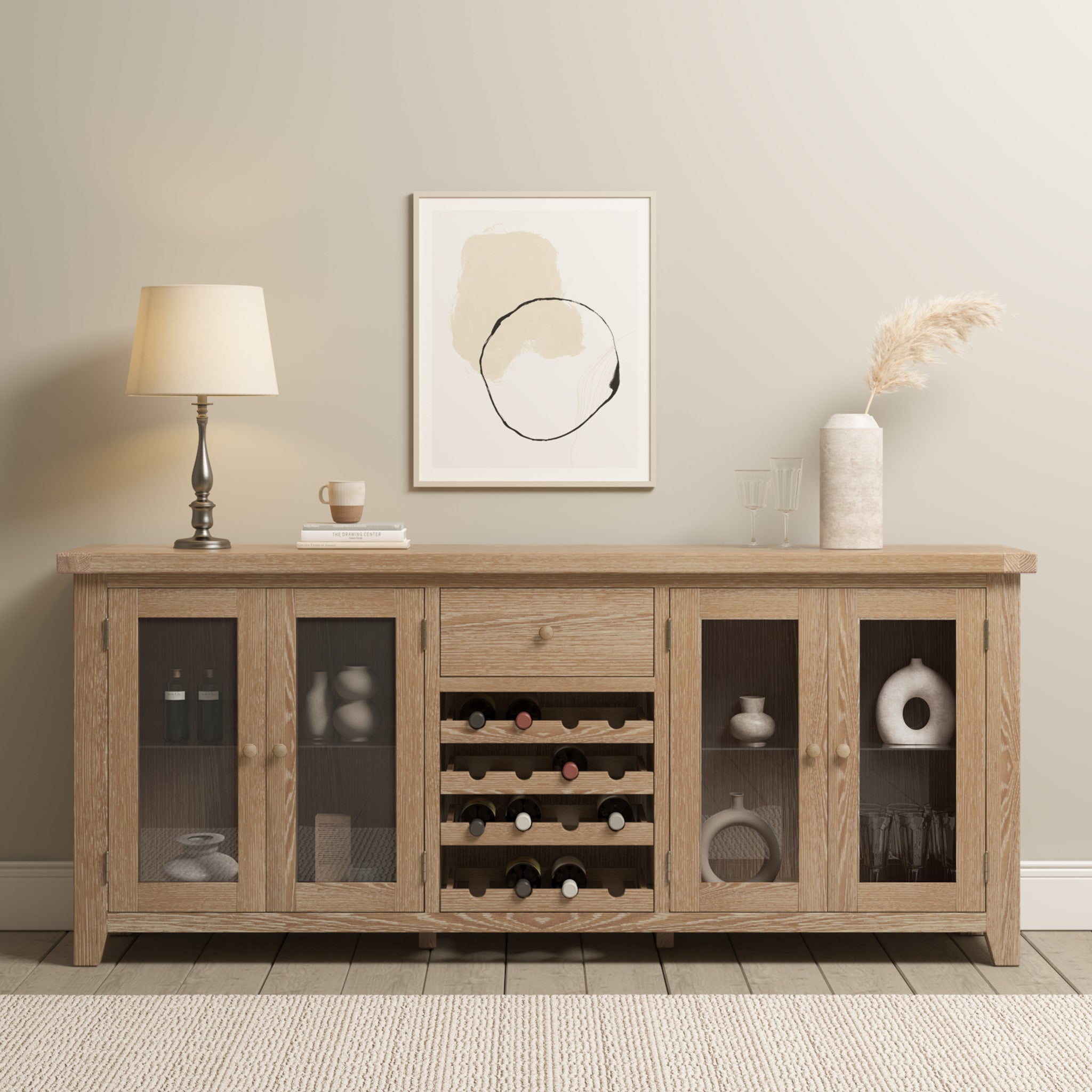 The Burford Grand Sideboard with Wine Rack in Natural Oak, elegantly decorated with a lamp, artwork, and other decorative pieces, stands against a softly hued wall.