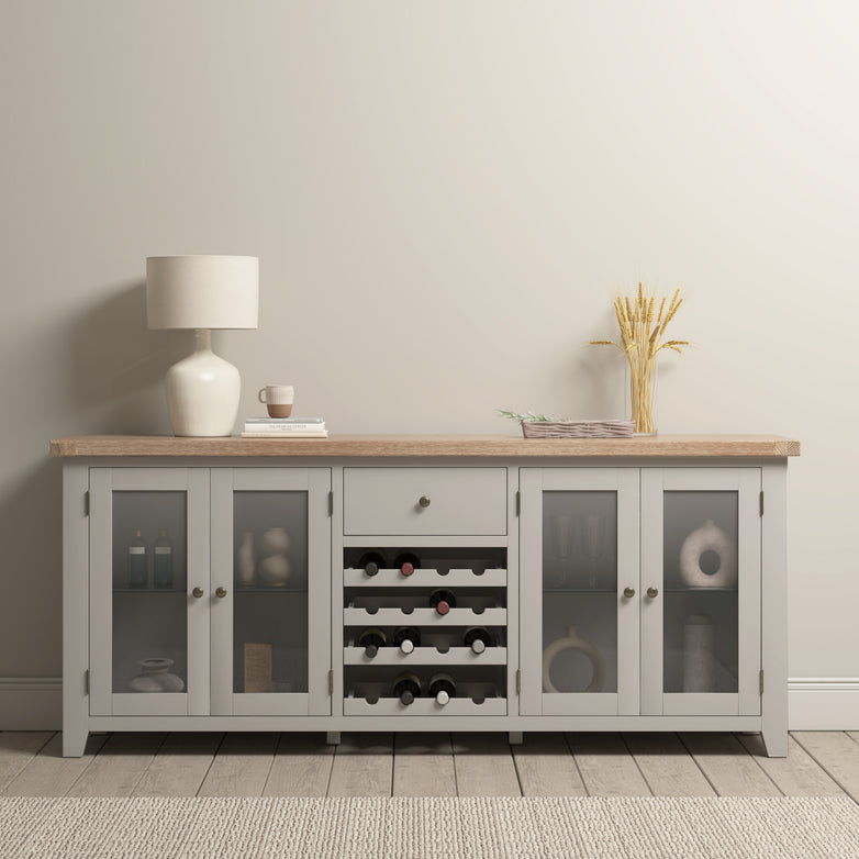 The Burford Grand Sideboard with Wine Rack in Pebble Grey beautifully displays a tabletop lamp, wine storage, ample space for various items, and decorations against a tranquil beige wall.