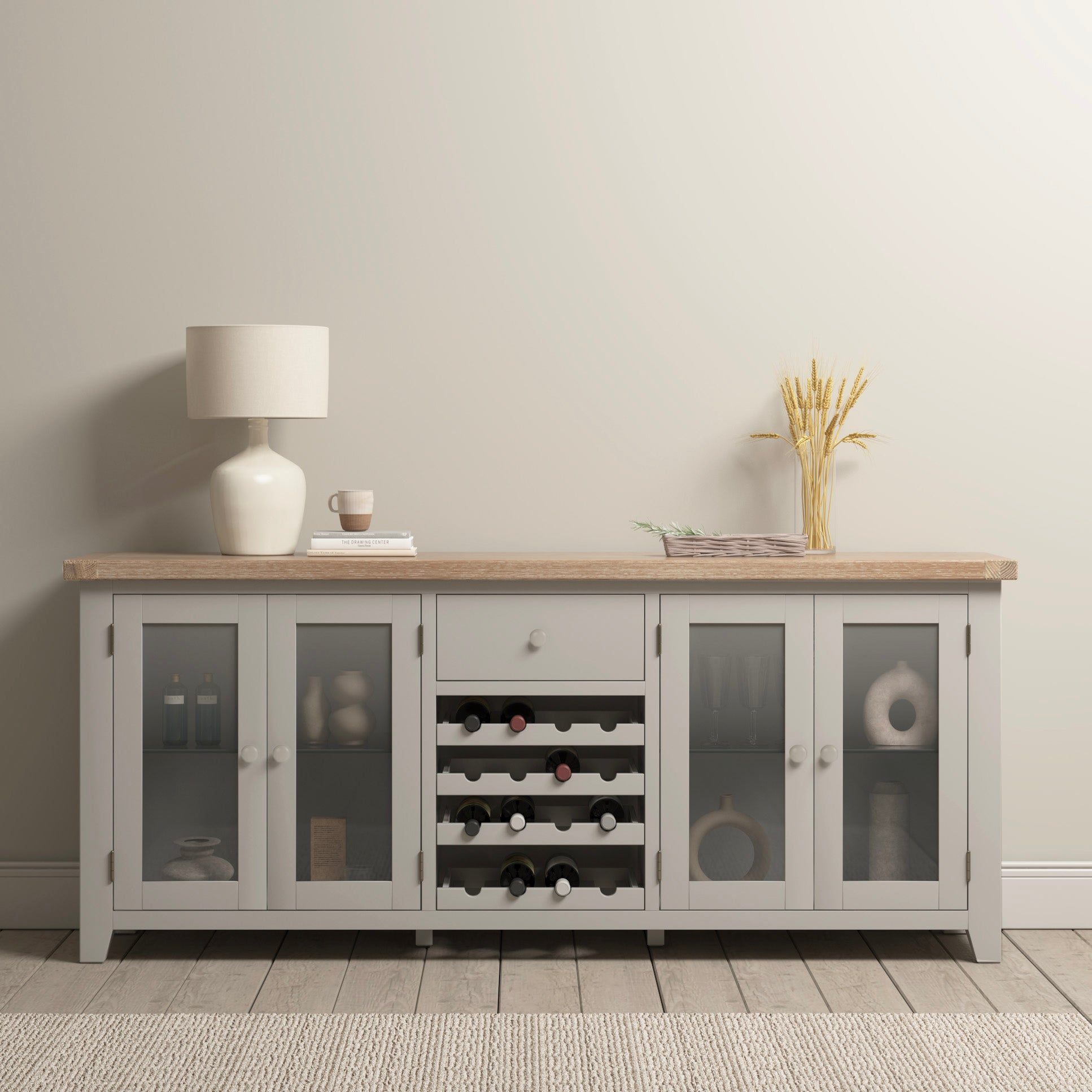 The Burford Grand Sideboard with Wine Rack in Pebble Grey gracefully presents its glass doors displaying fine china, features a built-in wine rack for treasured vintages, and is beautifully adorned with a lamp, cup, and vase filled with wheat on the wooden floor against a neutral wall.