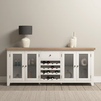 The Burford Grand Sideboard with Wine Rack in Warm White, crafted from natural oak, features elegant glass doors. Complementing decorative vases rest atop this exquisite piece, perfectly set against a neutral wall.