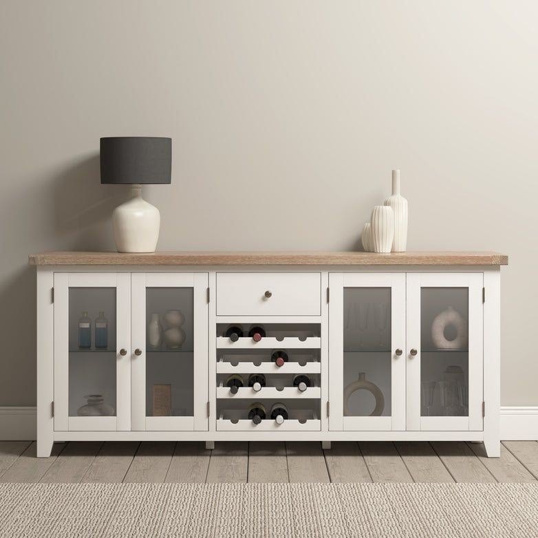 The Burford Grand Sideboard with Wine Rack in Warm White, crafted from natural oak, features elegant glass doors. Complementing decorative vases rest atop this exquisite piece, perfectly set against a neutral wall.