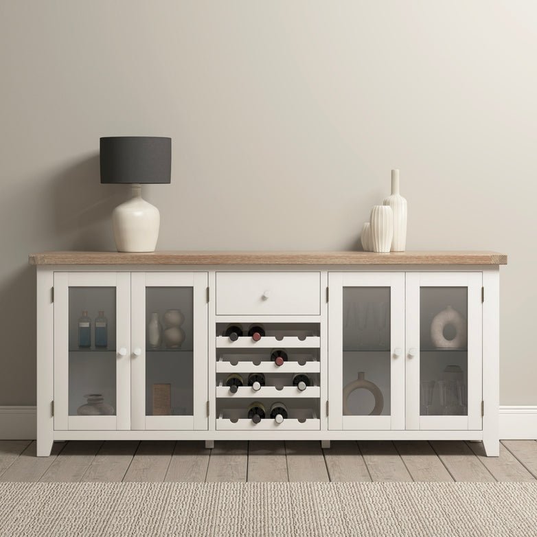 The Burford Grand Sideboard with Wine Rack in Warm White, featuring a natural oak top, includes glass doors and is tastefully accented by a black lamp and white vases, creating a harmonious look with the wooden floor.