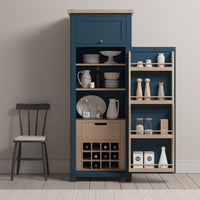 The Burford Single Larder Unit in Navy Blue features open shelves that beautifully display dishes, wine bottles, and condiments, all highlighted by a natural oak finish. A chair is placed on the left, enhancing the seamless blend of style and functionality in this inviting space.