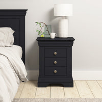 The Chalbury 3 Drawer Bedside Table in Dusky Black, made from solid oak, showcases drawers and holds a lamp, mug, and vase with branches. It is stylishly situated next to a bed on a light wood floor.