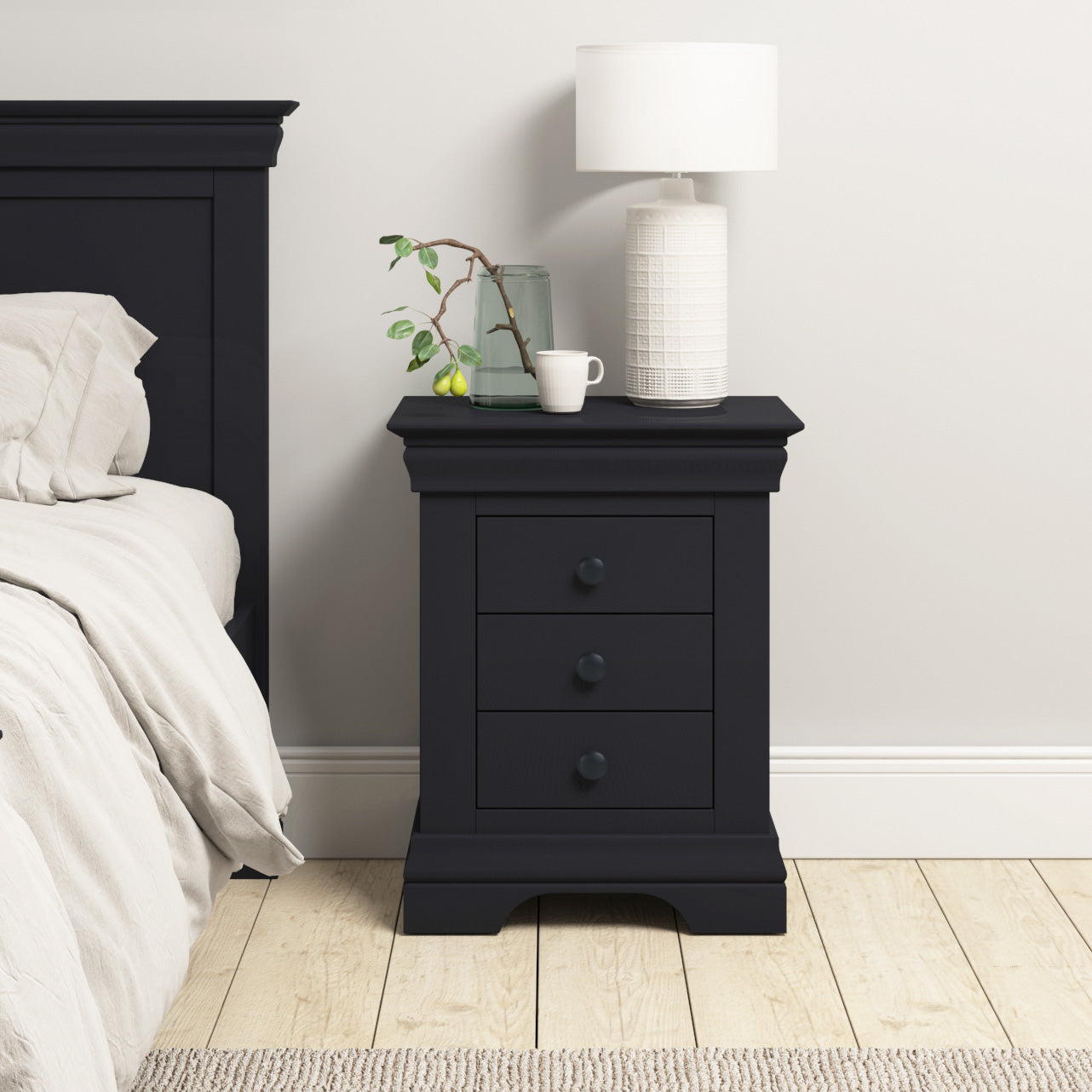 Introducing the Chalbury 3 Drawer Bedside Table in Dusky Black, a stunning creation made from solid oak. Paired with an elegant white lamp, a small plant, and a cozy mug, it beautifully complements light bedding and wooden floors, adding charm beside your bed.