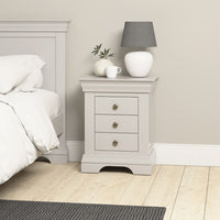 A Chalbury 3 Drawer Bedside Table in Pebble Grey, adorned with a lamp, potted plant, and cup, is placed beside the bed in a bright room.