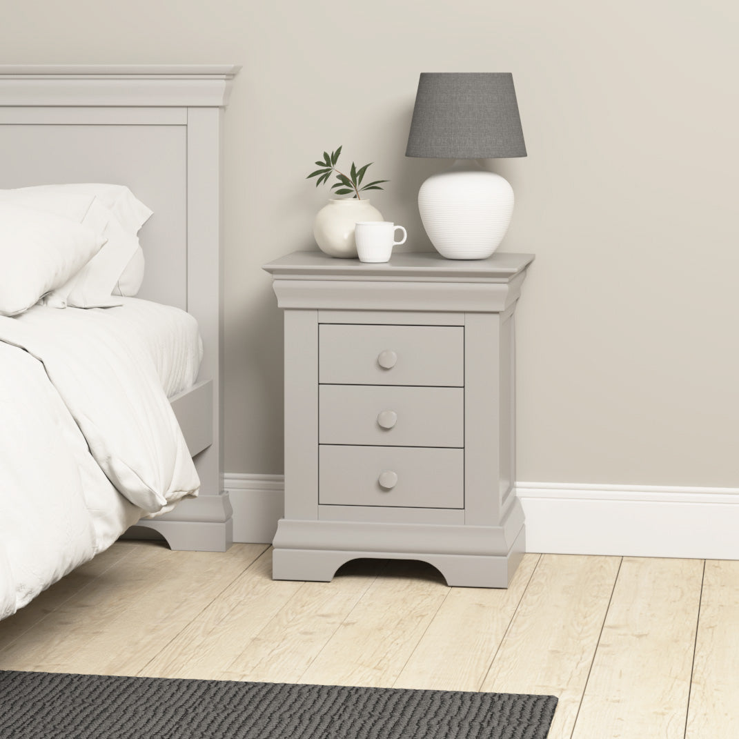 Chalbury 3 Drawer Bedside Table in Pebble Grey featuring a lamp, a plant, and a mug beside a bed in an inviting bedroom setting.