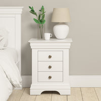 The Chalbury 3 Drawer Bedside Table in Warm White adds elegance beside the bed in a brightly lit room. It is adorned with stylish bedroom décor—a plant, a cup, and a lamp—offering both charm and functionality.