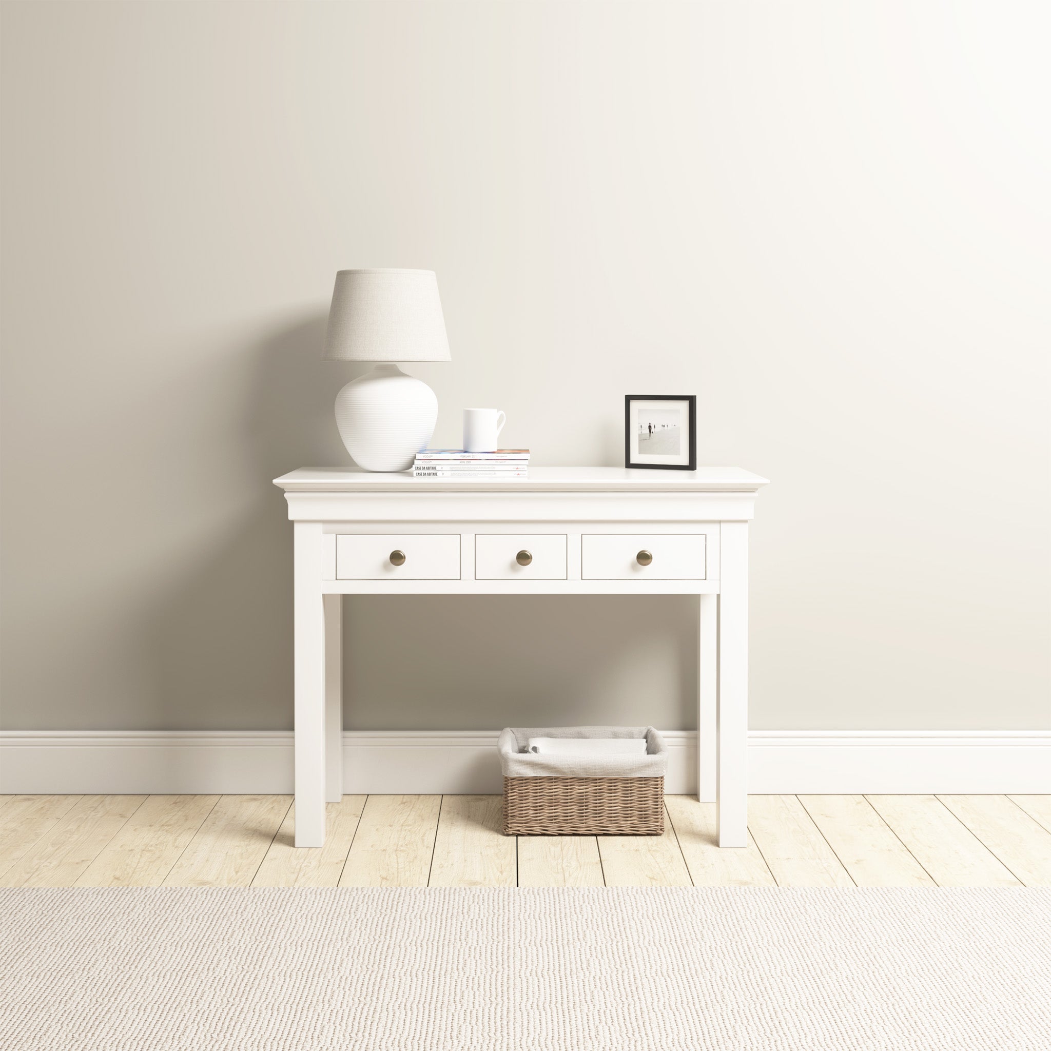 The Chalbury Console Desk in Warm White exudes modern elegance, adorned with a lamp, books, and a photo frame. The wicker basket below complements the minimalistic room beautifully.