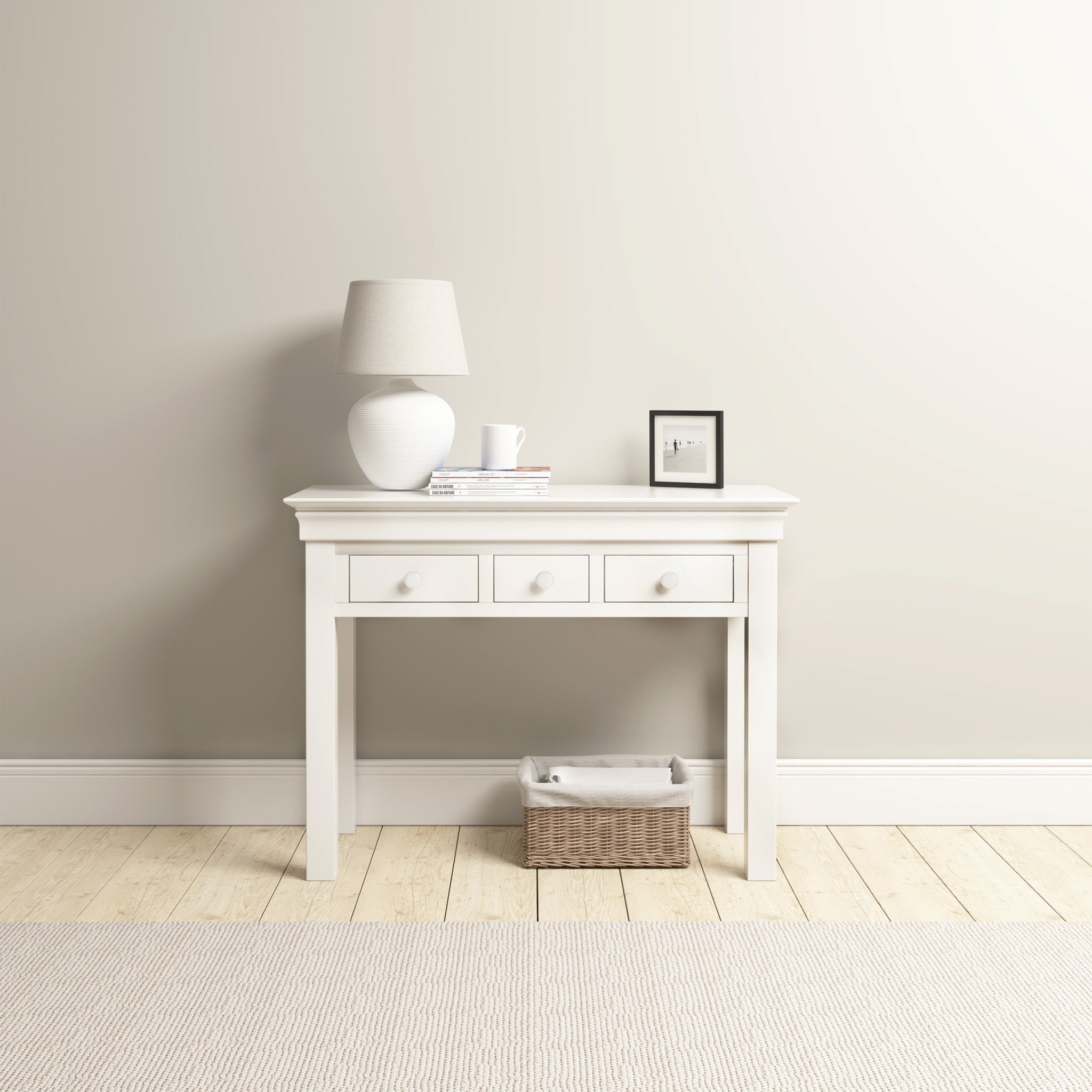 The Chalbury Console Desk in Warm White embodies a sense of modern elegance, highlighted by its sleek design that includes drawers, a lamp, books, and a framed picture. Below it sits a wicker basket that fits naturally beneath the desk, while everything is set beautifully on the polished wooden floor.