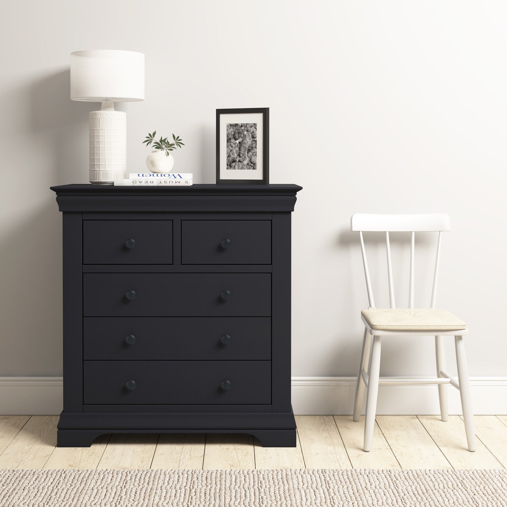 The Chalbury 2 Over 3 Chest of Drawers in Dusky Black, paired with a lamp and photo, complements a white chair on a wooden floor with a light rug, showcasing modern design.