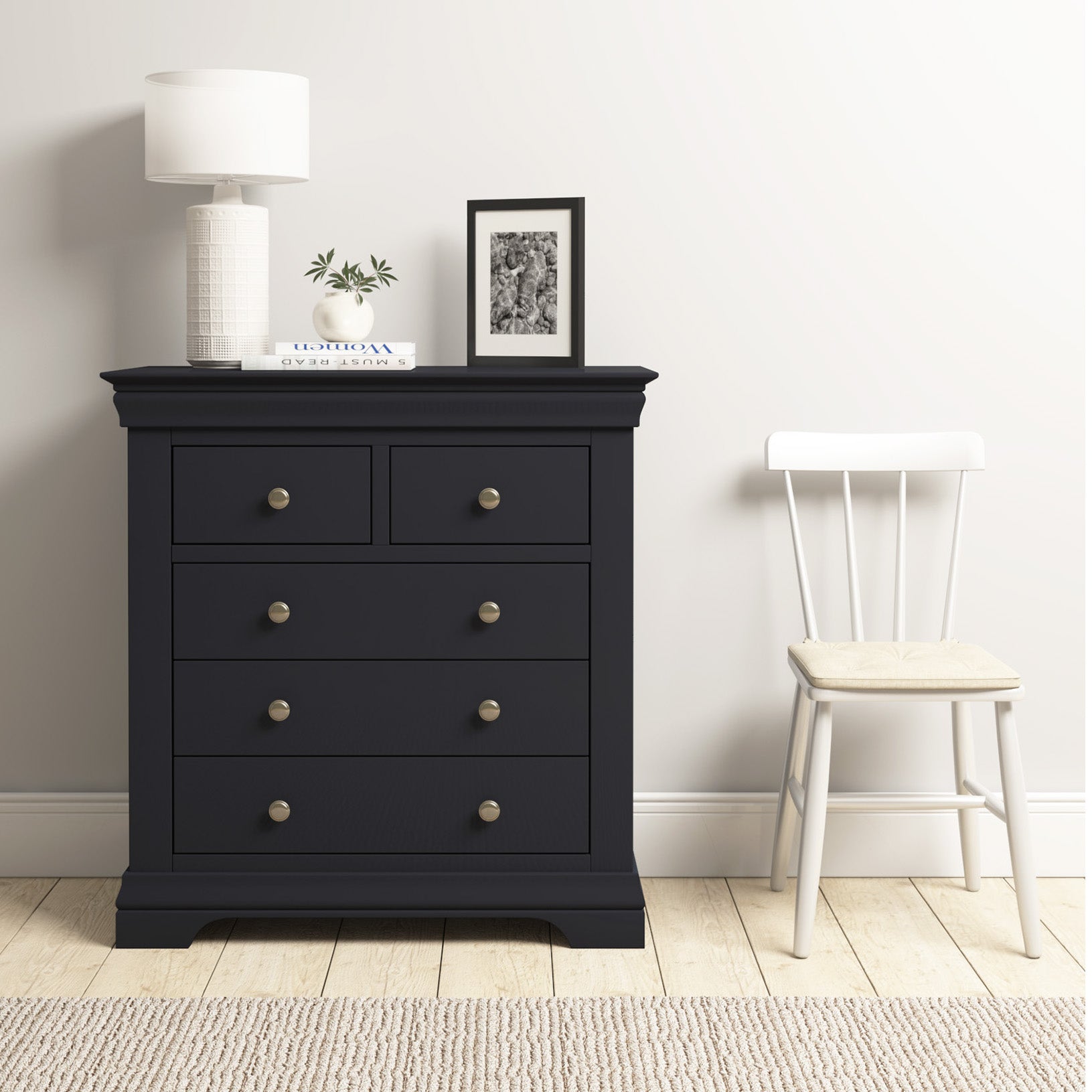 A Chalbury 2 Over 3 Chest of Drawers in Dusky Black, decorated with a lamp, plant, and photo frame, sits stylishly next to a white chair on the wooden floor.