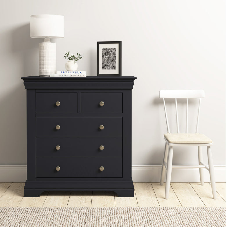 A Chalbury 2 Over 3 Chest of Drawers in Dusky Black, decorated with a lamp, plant, and photo frame, sits stylishly next to a white chair on the wooden floor.