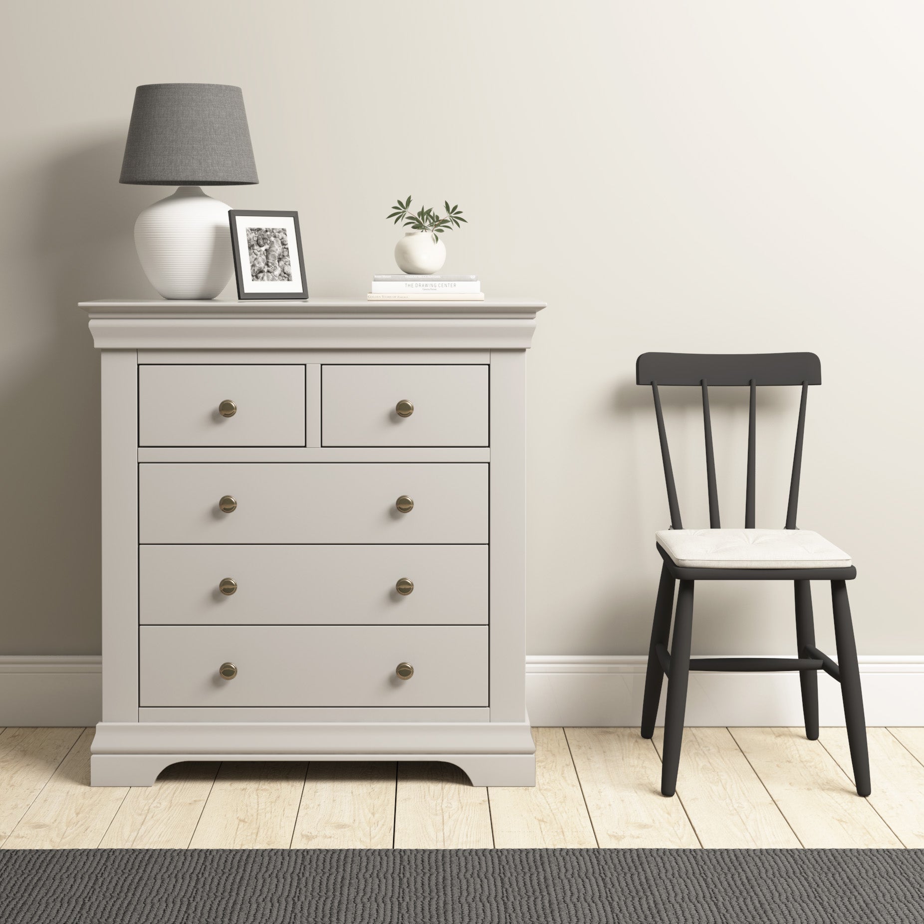 A modern arrangement showcases the Chalbury 2 Over 3 Chest of Drawers in Pebble Grey, accompanied by a lamp, framed photo, and plant, positioned beside a black chair on a wooden floor.