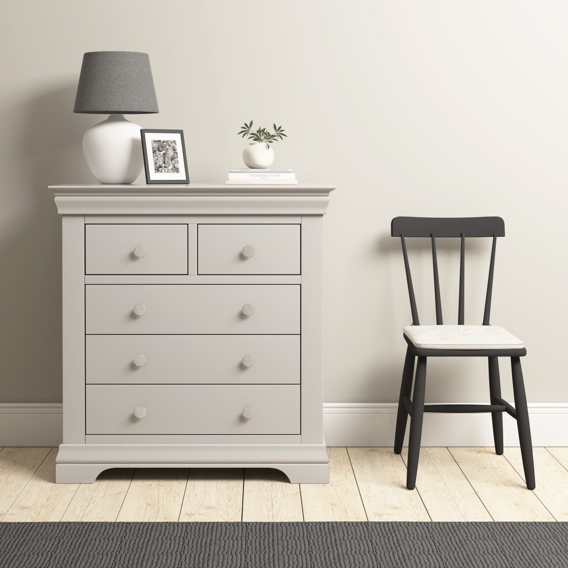 A Chalbury 2 Over 3 Chest of Drawers in Pebble Grey, adorned with a sleek lamp and stylish decor, stands next to a black chair on a wood floor, perfectly embodying contemporary design.