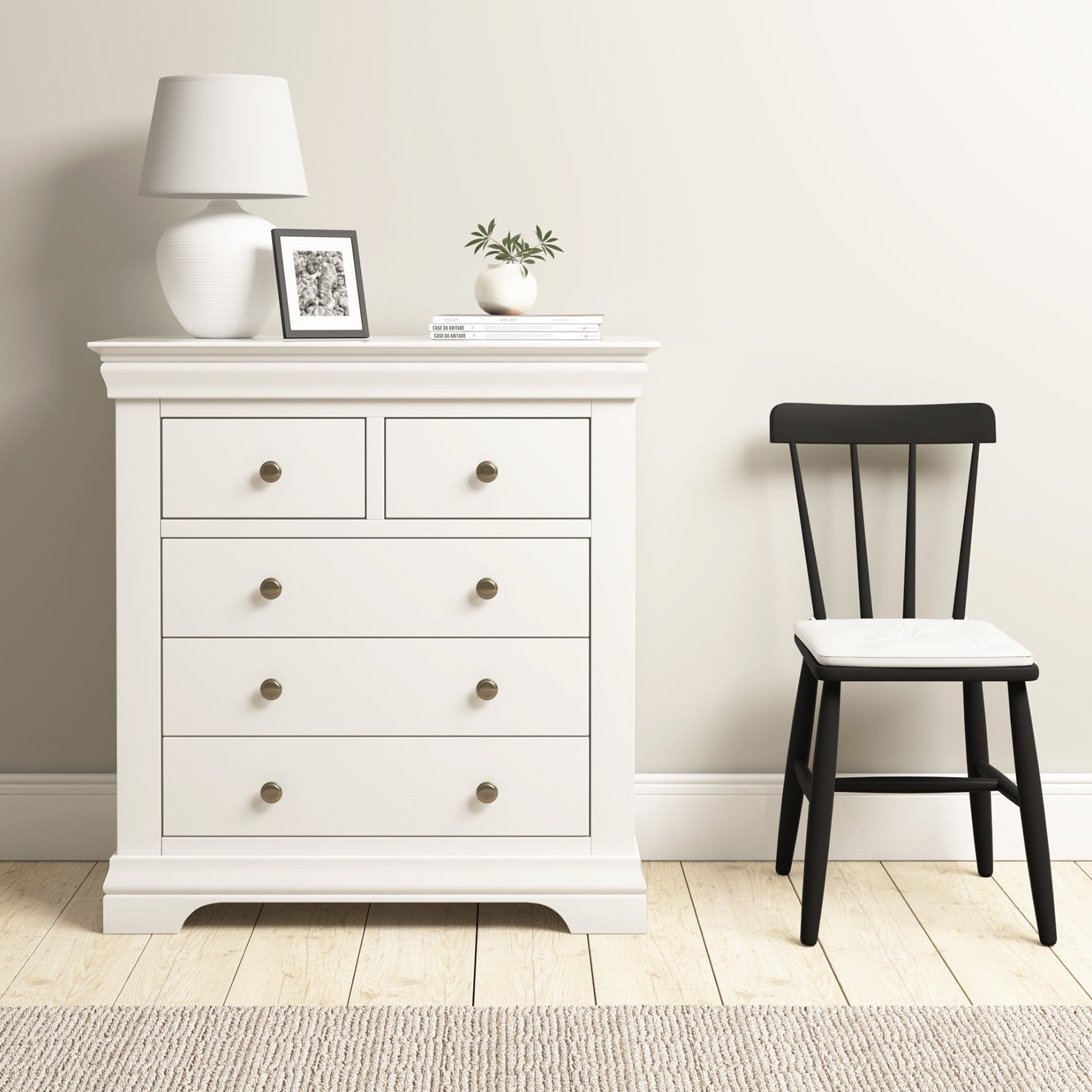 The Chalbury 2 Over 3 Chest of Drawers in Warm White, with modern design and ample storage, sits elegantly with a lamp and decor beside a black chair on a wooden floor.