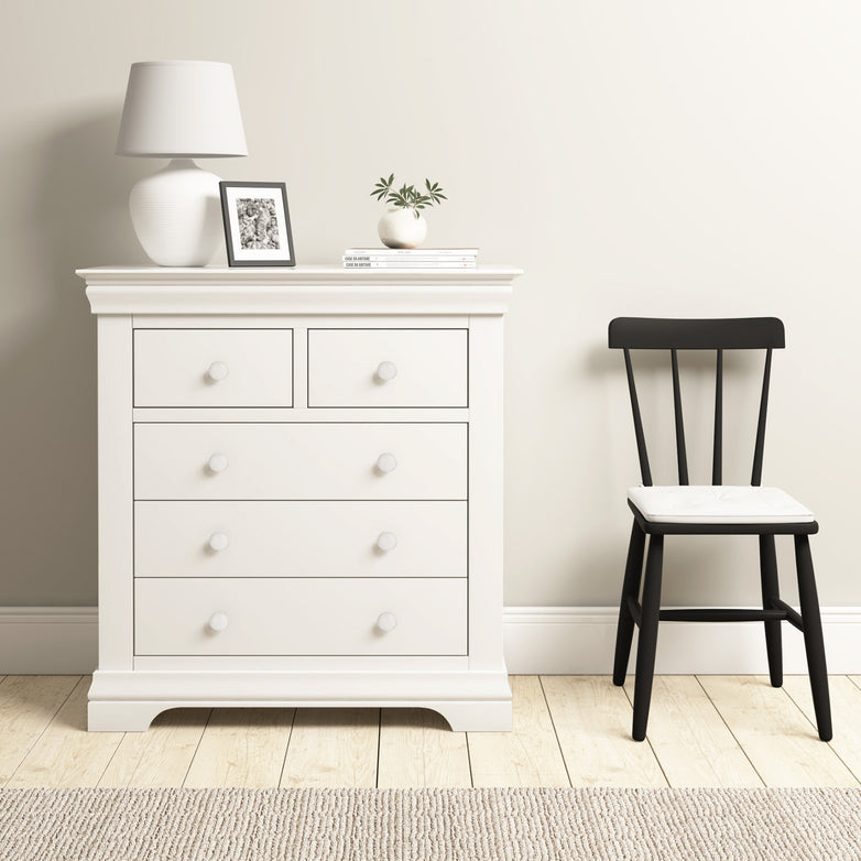 The warm white Chalbury 2 Over 3 Chest of Drawers blends modern design and ample storage, elegantly complementing a black chair on the light wood floor, with decor items atop adding a touch of sophistication.