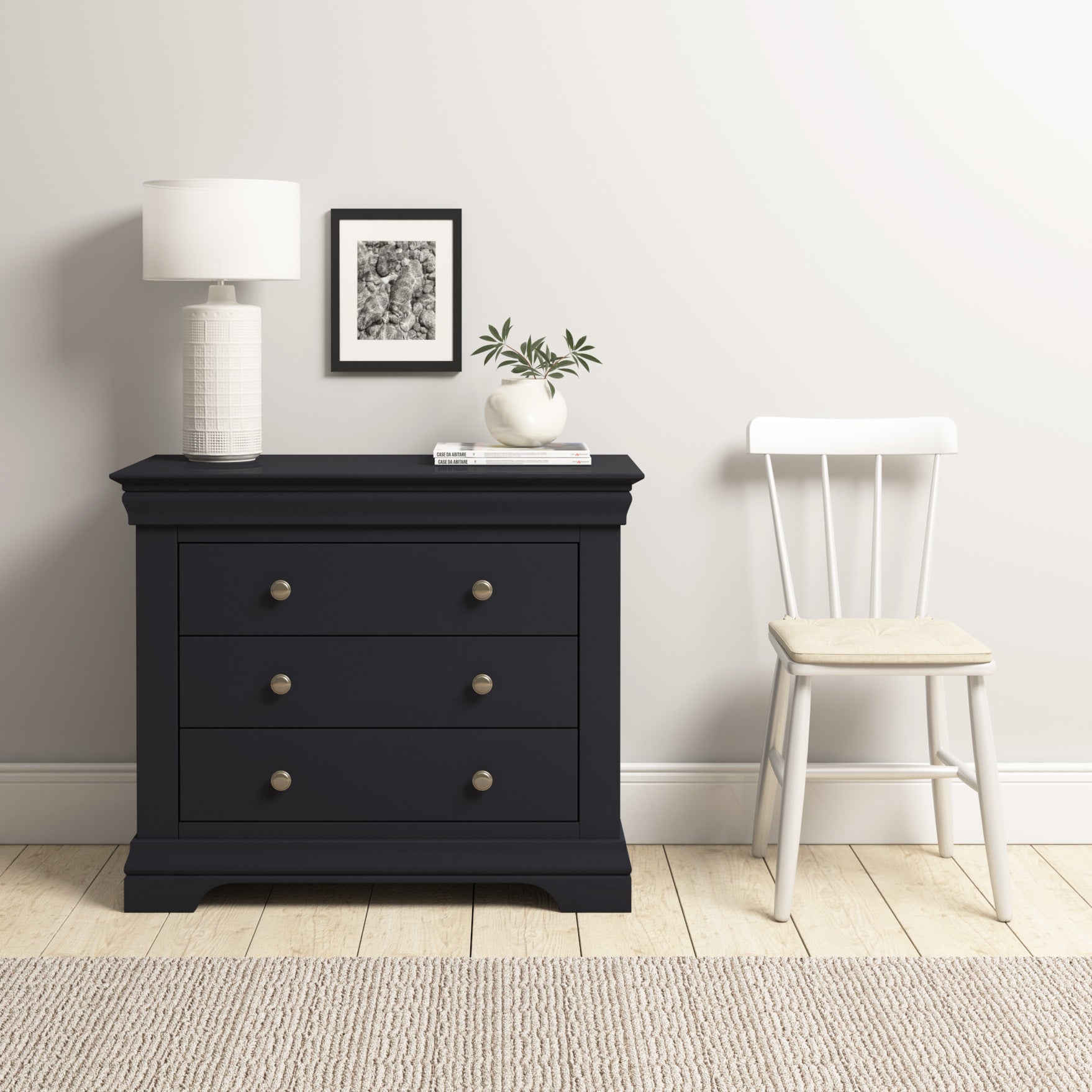 A Chalbury 3 Drawer Chest in Dusky Black stylishly supports a lamp, framed picture, and plant beside a white chair on a light wood floor.