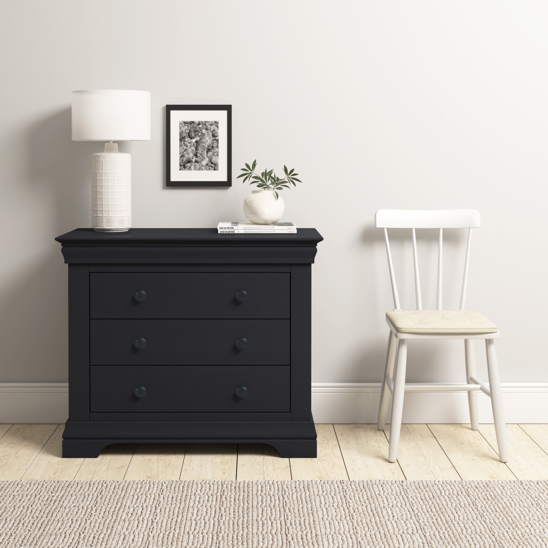 The Chalbury 3 Drawer Chest in Dusky Black, adorned with a lamp, framed photo, and plant, perfectly complements the white chair on the light wood floor with a rug. Its modern design adds an elegant touch to any room.