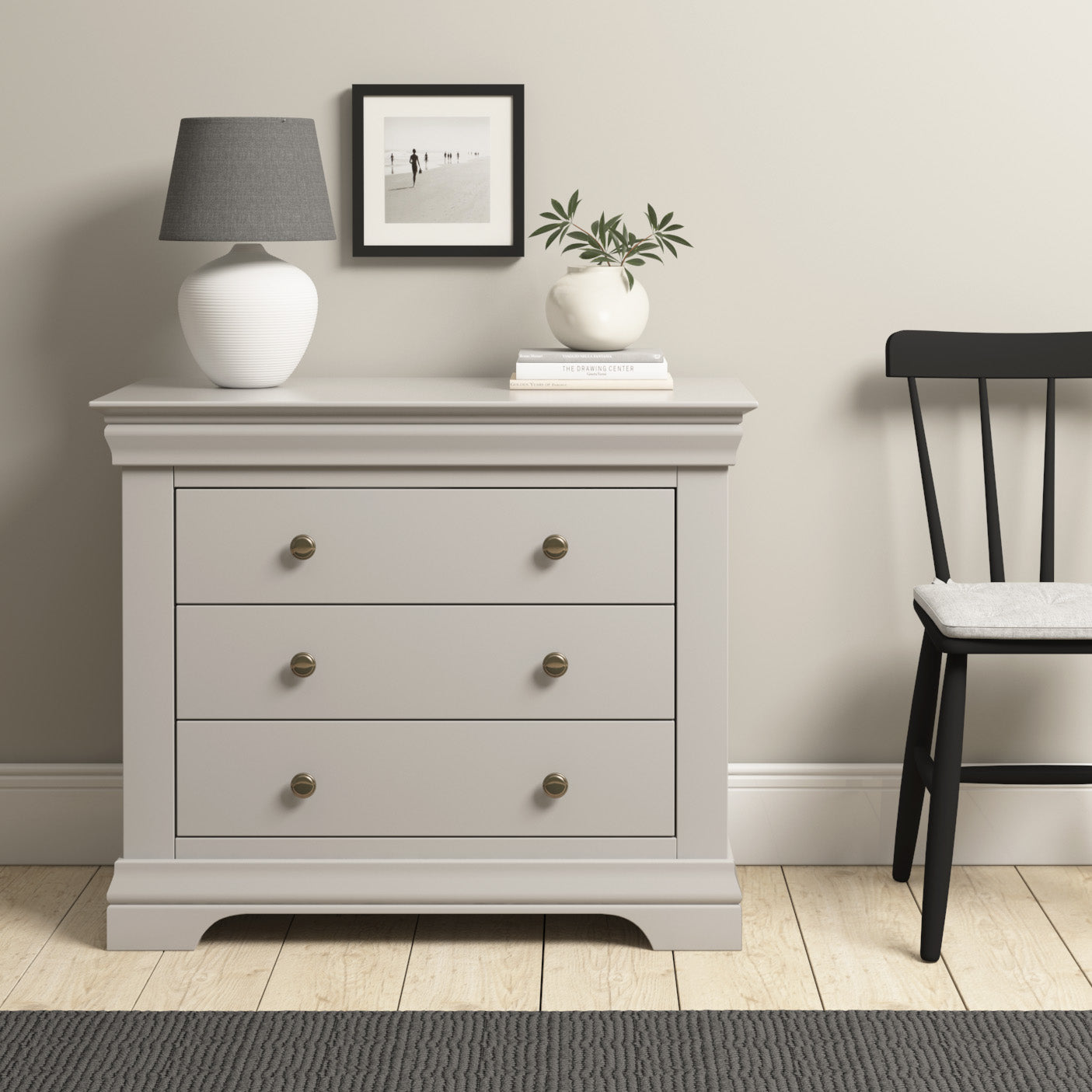 The Chalbury 3 Drawer Chest in Pebble Grey enhances the room's elegance with its display of a lamp, books, and a plant. Positioned beside a black chair on a wooden floor, this chic storage piece creates a cozy and organized nook.