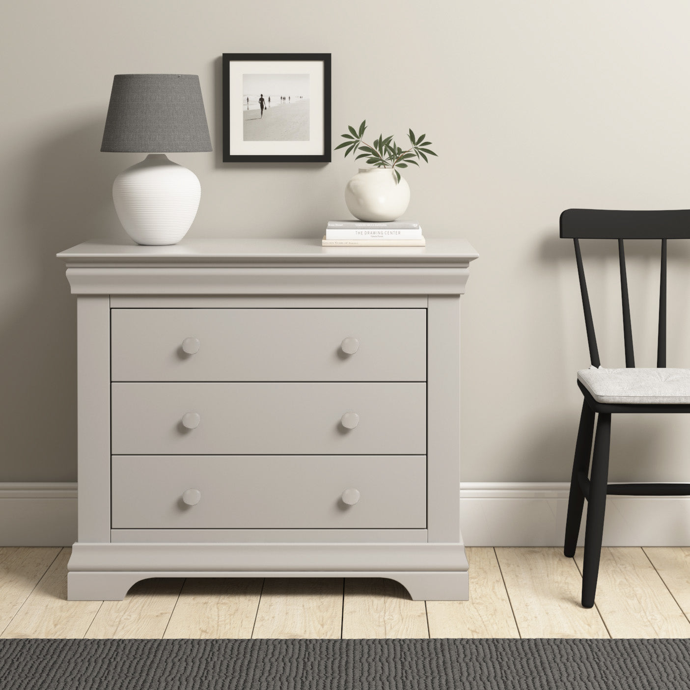 The Chalbury 3 Drawer Chest in Pebble Grey provides elegant storage with its three roomy drawers. Styled with a black chair beside it, a lamp, and a small plant in a vase on its surface, it complements the photo frame hanging above.