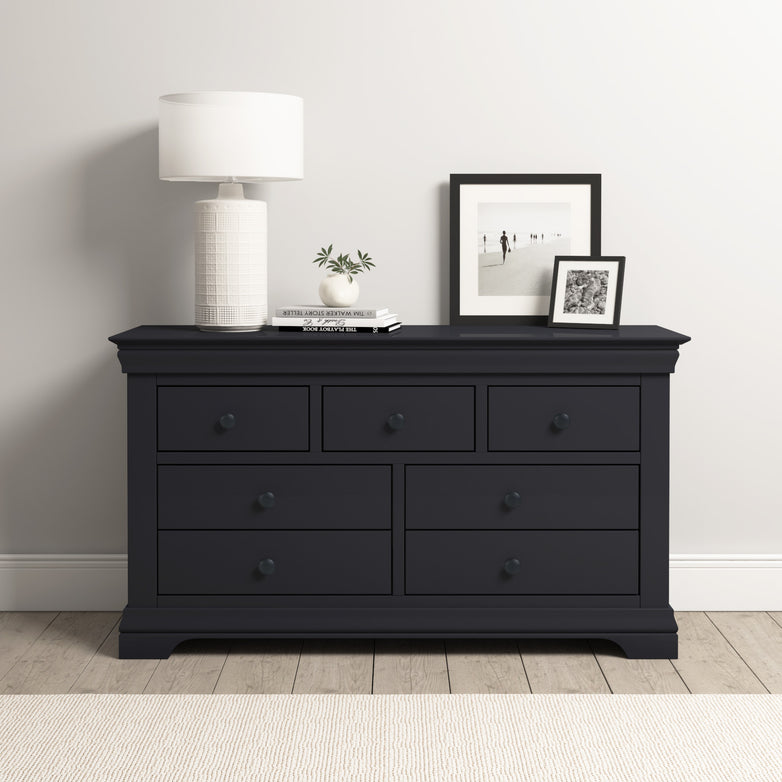 The minimalist room showcases a Chalbury 3 Over 4 Chest of Drawers in Dusky Black, elegantly topped with a lamp, books, and framed photos. Positioned against a crisp white wall, this chest seamlessly merges functionality with style.