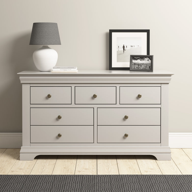 The Chalbury 3 Over 4 Chest of Drawers in Pebble Grey provides extensive storage with its seven drawers, accentuated by a matching gray lamp and black-and-white photographs, all beautifully arranged on a wooden floor.