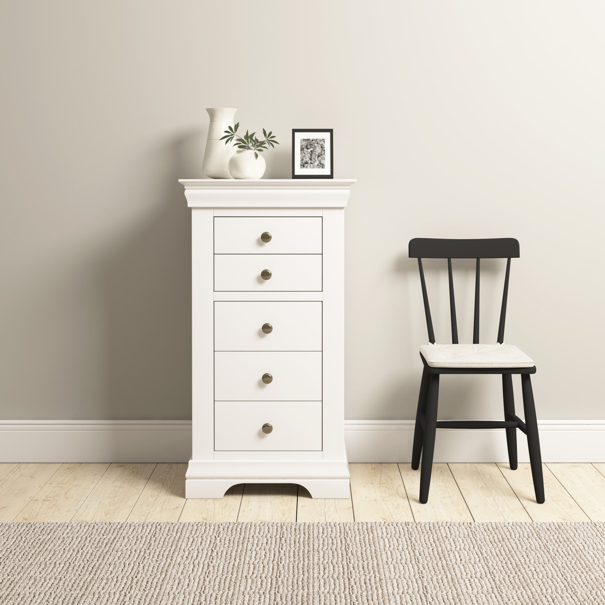 Chalbury 5 Chest of Drawers in Warm White, adorned with decor, stands beside a black chair on a light wooden floor against a beige wall, capturing the essence of elegance.