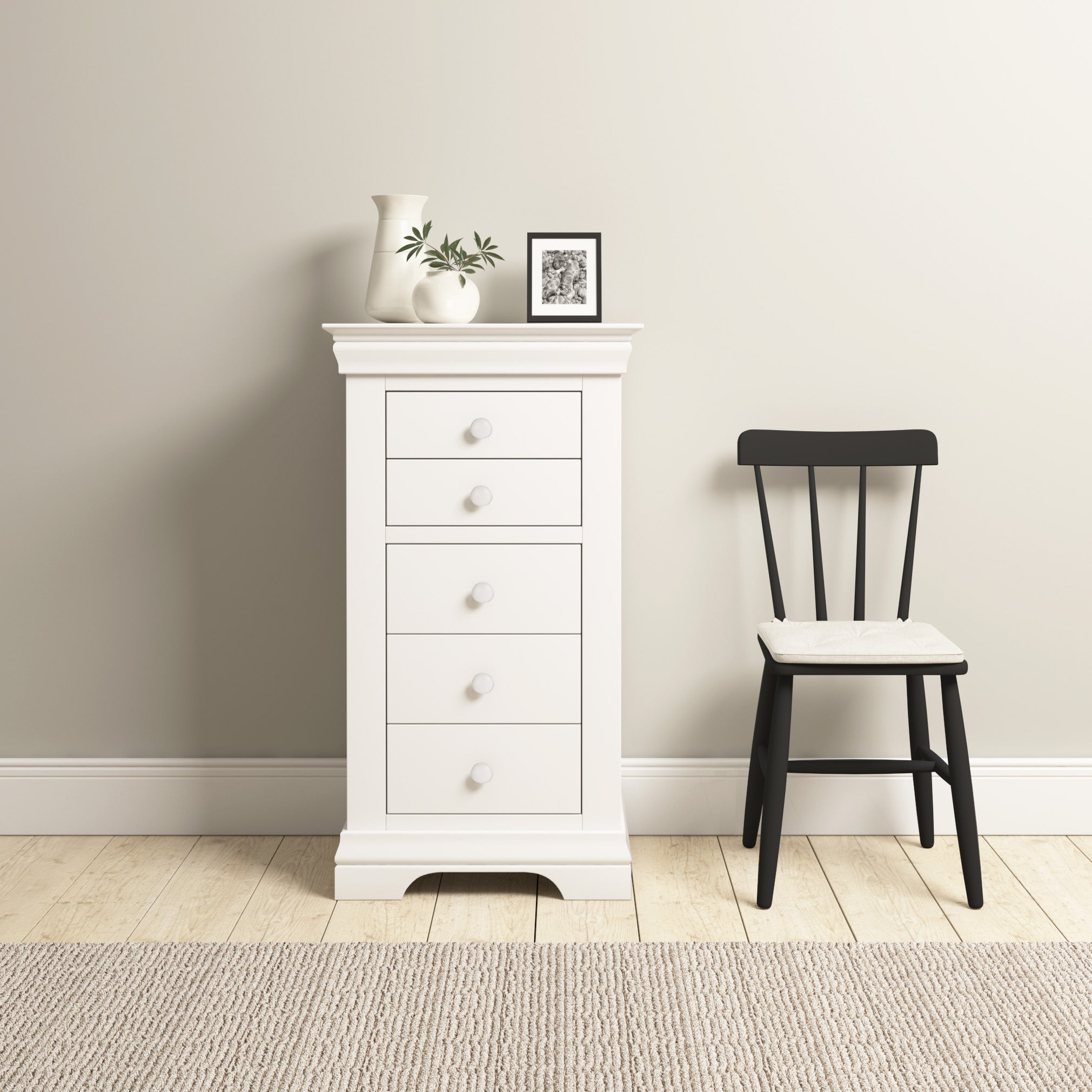 The Chalbury 5 Chest of Drawers in Warm White stands elegantly with its five spacious drawers beside a black chair and curated decor items against a light wall, adding charm to the cozy room.