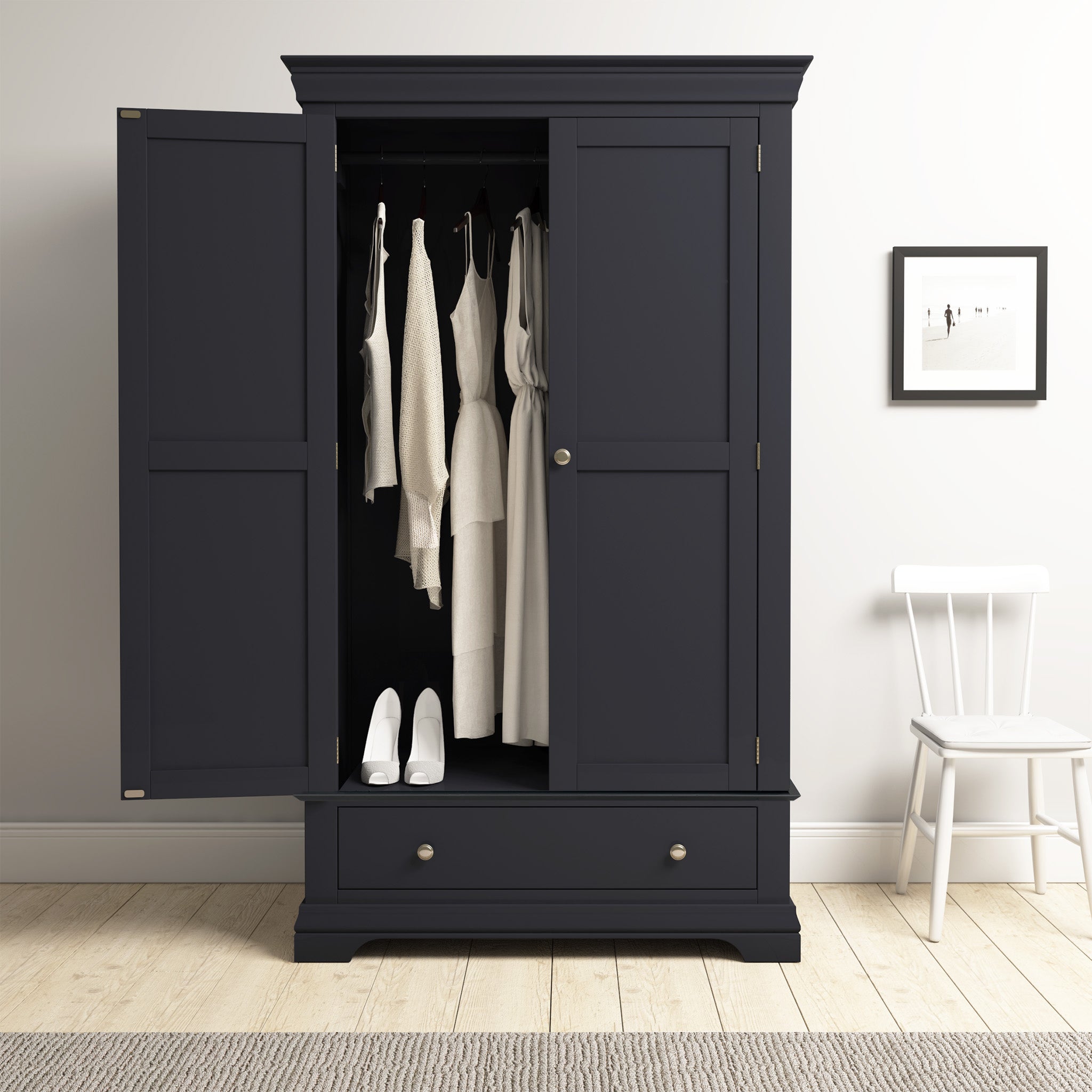 The Chalbury Double Wardrobe in Dusky Black offers ample storage for neatly organized white clothes and shoes, complemented by a nearby white chair and framed picture for a chic look.