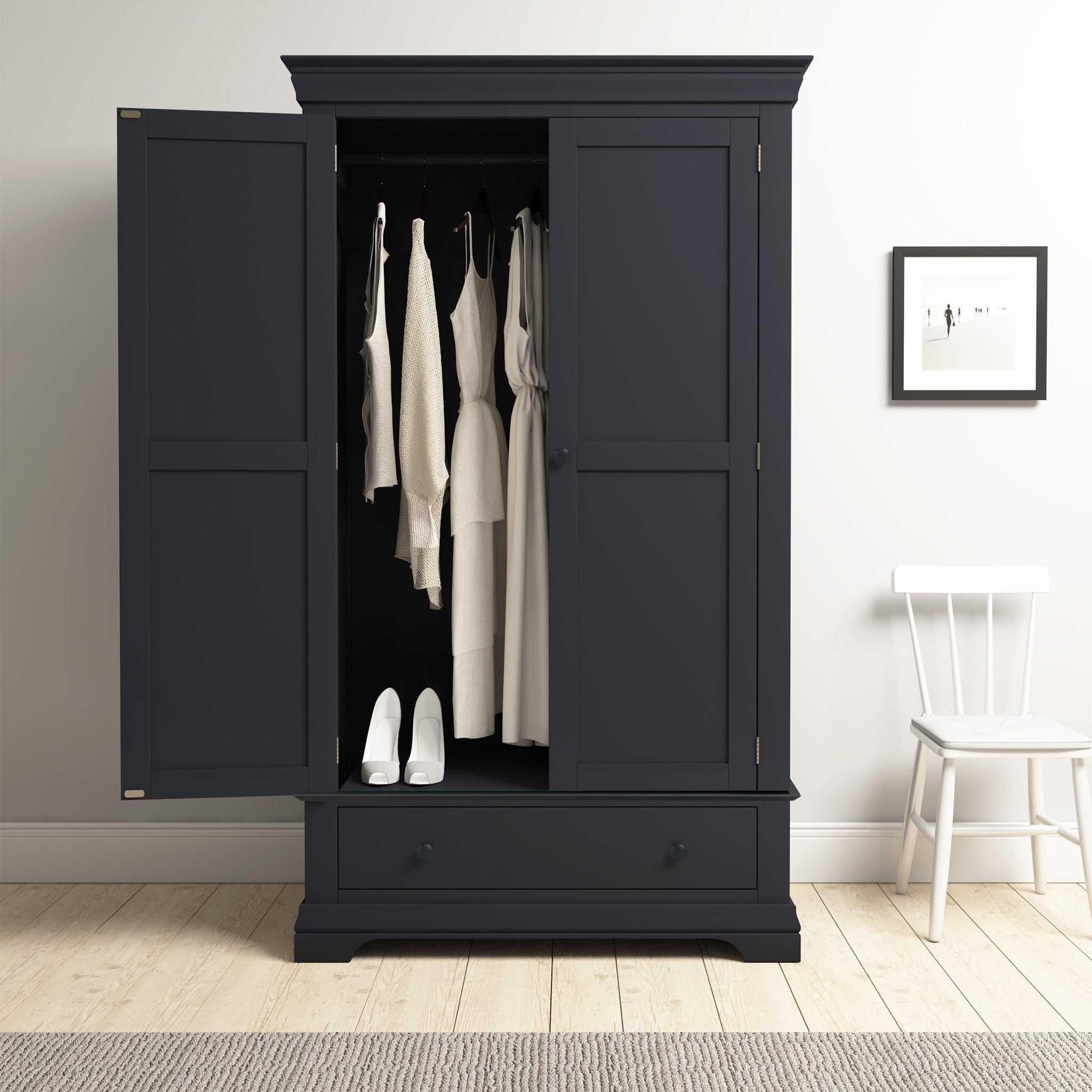 The Chalbury Double Wardrobe in Dusky Black features open doors showcasing white clothes and shoes within its ample storage, paired with a white chair and framed art on display.