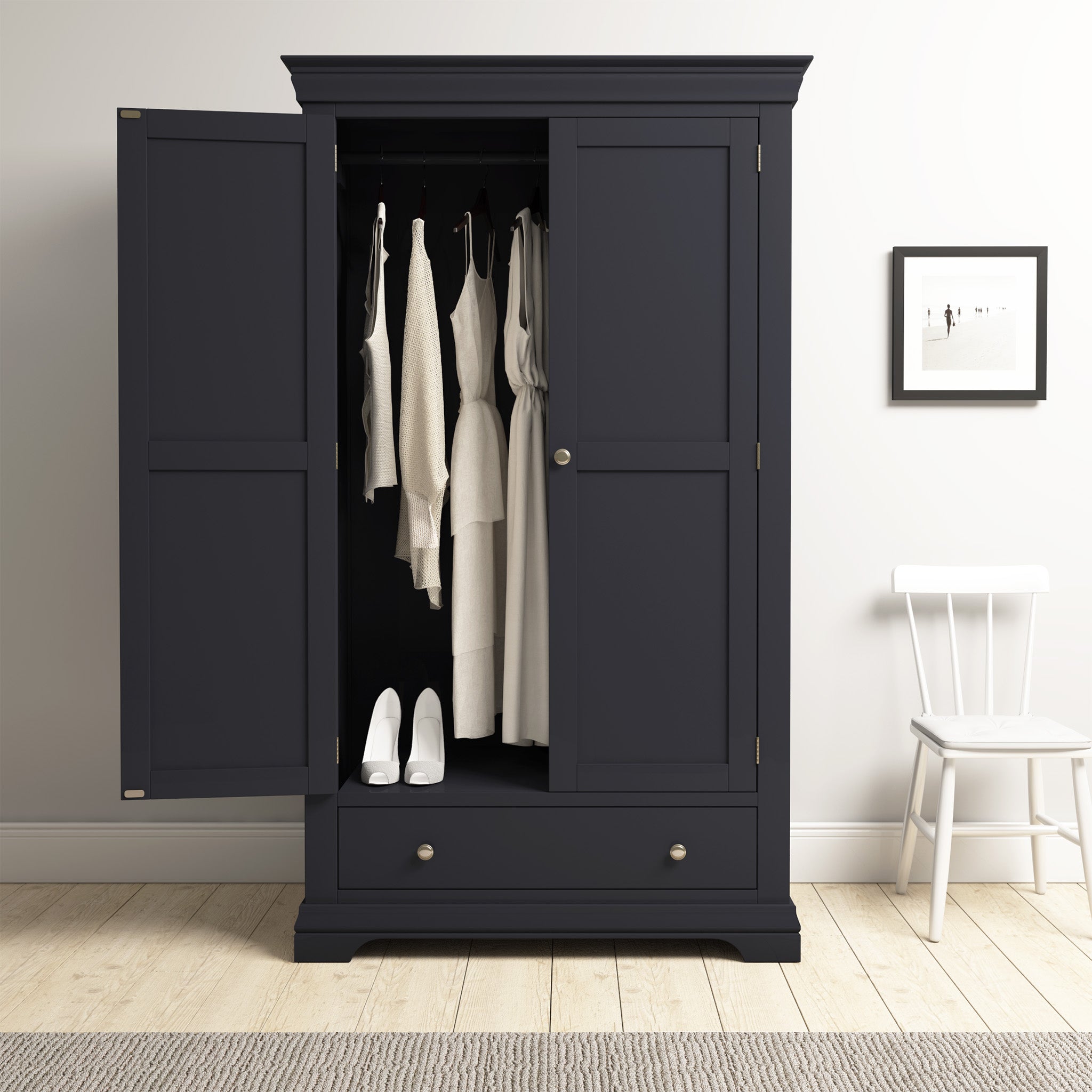 The Chalbury Double Wardrobe in Dusky Black stands proudly as a centerpiece of bedroom décor, its open doors revealing an elegant collection of white dresses and shoes inside, complemented perfectly by a nearby white chair and framed artwork.