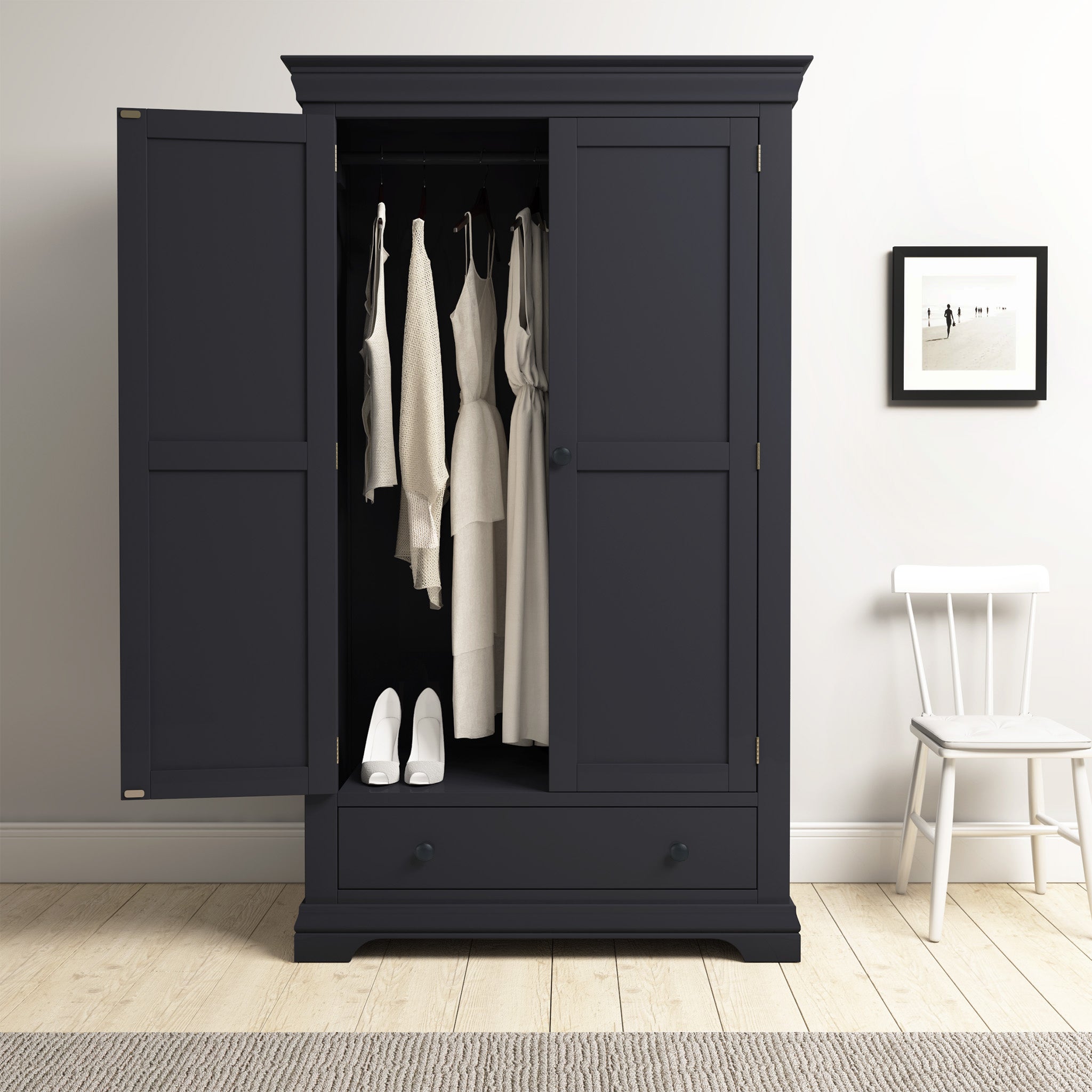 The Chalbury Double Wardrobe in Dusky Black stands with its doors open, showcasing neatly organized white clothing and footwear. Beside it, a white chair enhances the arrangement, while a framed picture adds a touch of charm to the bedroom decor.