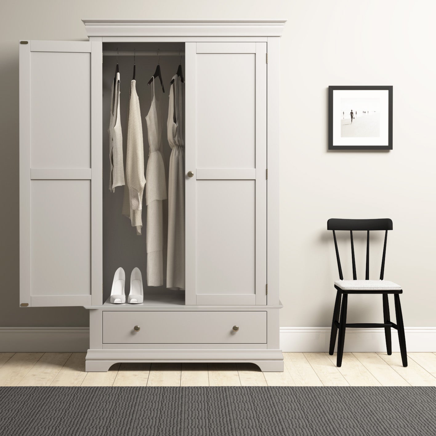 The Chalbury Double Wardrobe in Pebble Grey showcases a neat array of white clothes and shoes, while a black chair next to it enhances the modern look alongside a framed picture on the light wall.