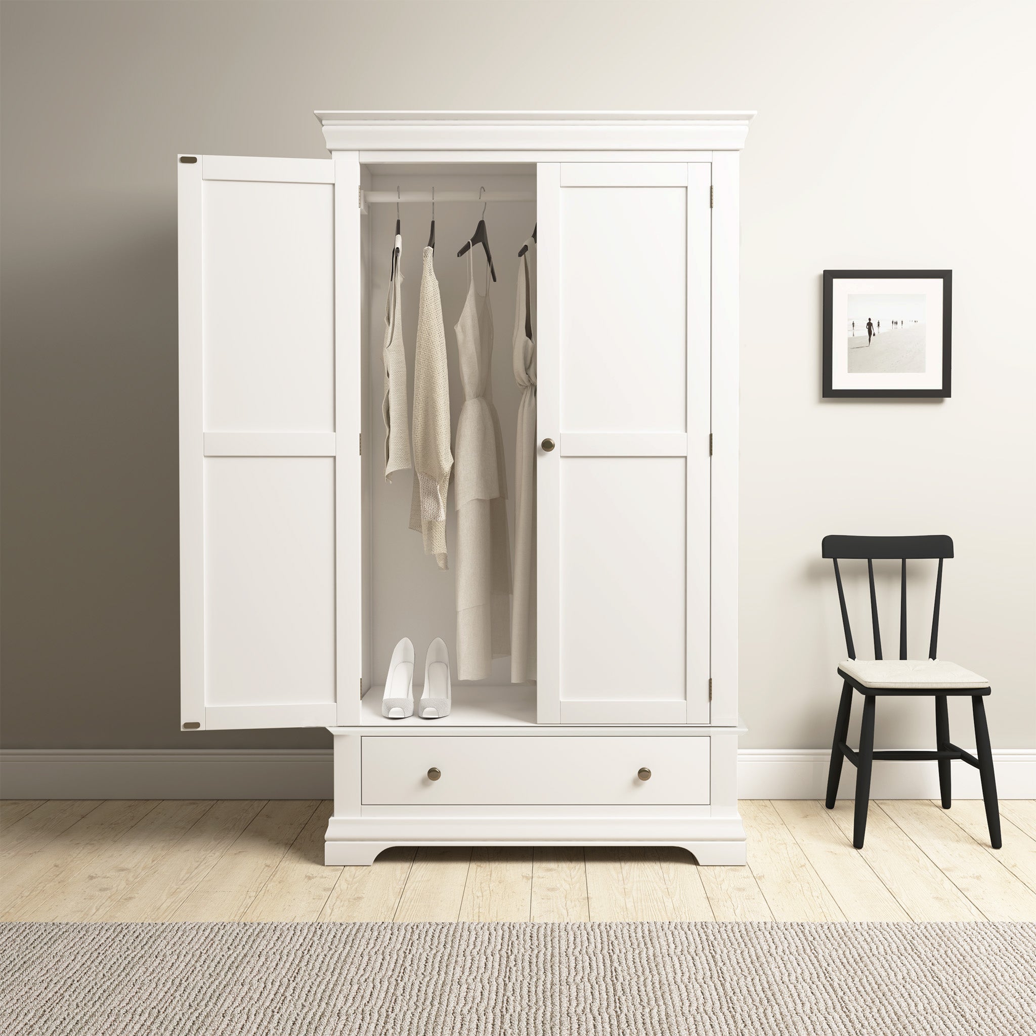 The Chalbury Double Wardrobe in Warm White, with its contemporary design, elegantly stores clothes and shoes. It complements a minimalist room featuring a chair and framed wall picture for a harmonious and stylish ambiance.