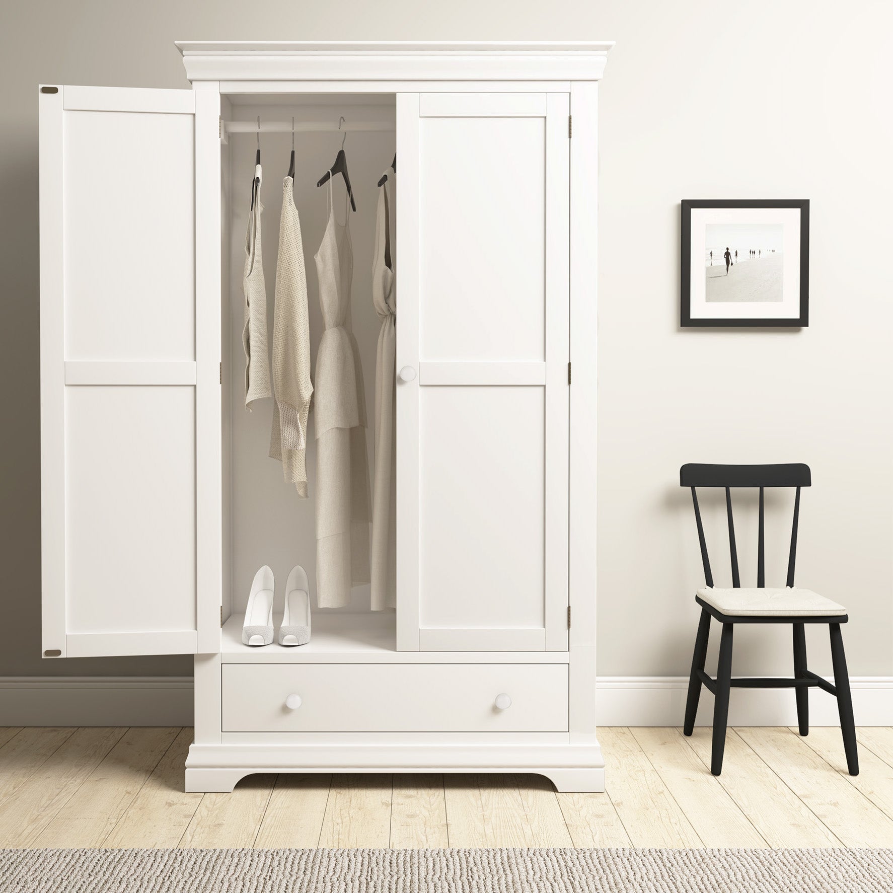 The Chalbury Double Wardrobe in Warm White provides plenty of space for neutral clothes and shoes, pairing perfectly with a black chair. A framed photo adds personalization to this tidy area.