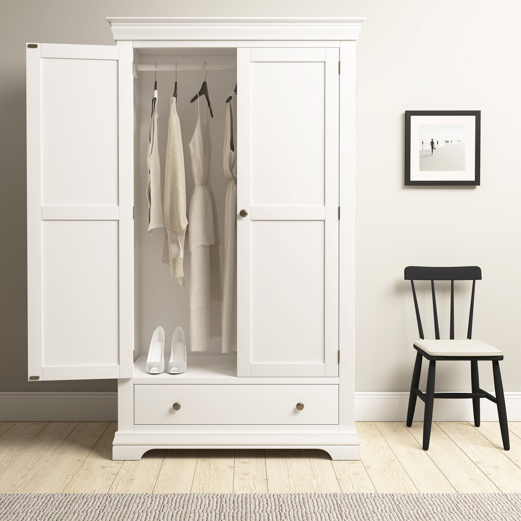 The Chalbury Double Wardrobe in Warm White opens to spacious storage for clothes and shoes, accompanied by a black chair and a decorative picture frame for an elegant, artistic touch.