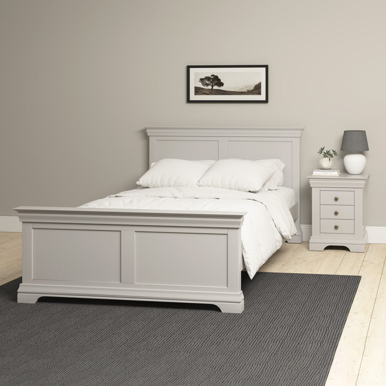 The minimalist bedroom features a high-end design with the Chalbury 4ft 6" Double Bed Frame in Pebble Grey, accompanied by an understated nightstand, a stylish lamp, and elegant wall art against a beige backdrop.