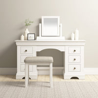 The Chalbury Dressing Table with Drawers in Warm White has six drawers, adorned with small plants and framed photos on top, and comes with a cushioned bench.