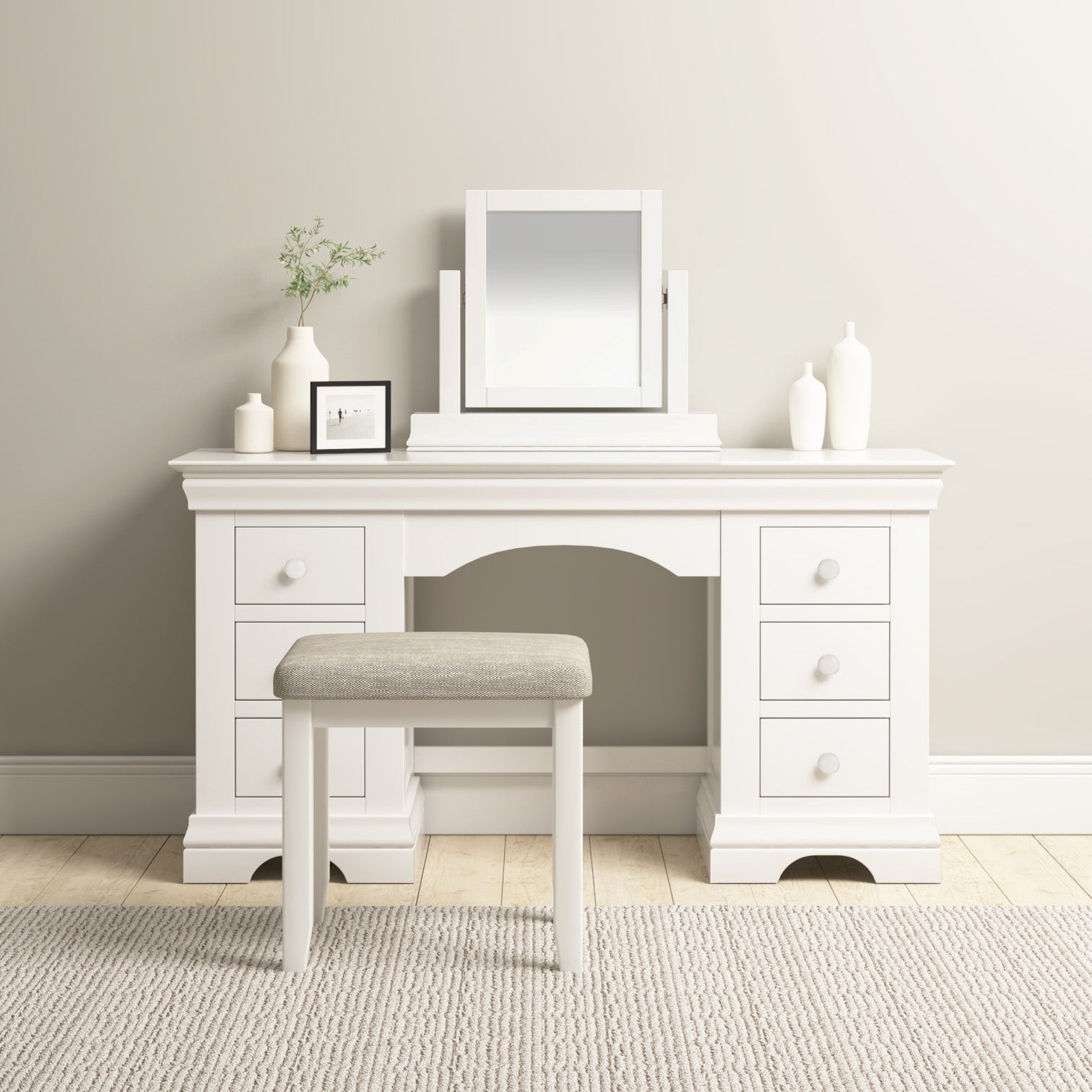 The Chalbury Dressing Table with Drawers in Warm White gracefully sits on the light wooden floor, featuring an accompanying stool and decorative vases. A mirror completes its enchanting presence.