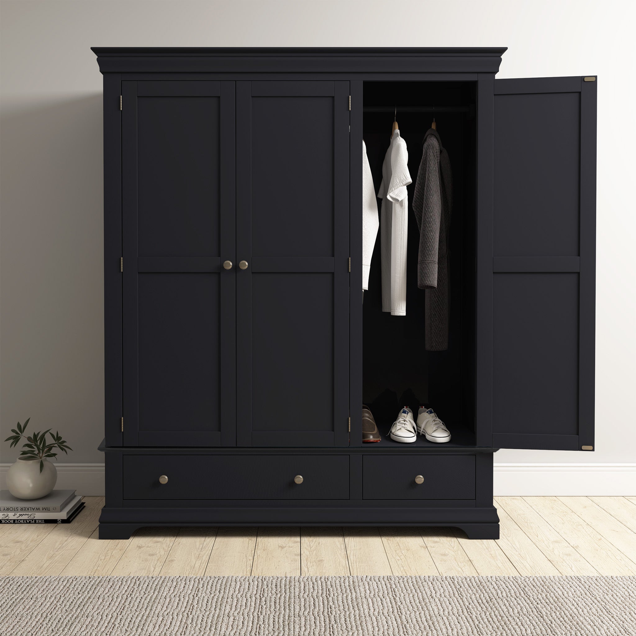 Against a white wall, the Chalbury Triple Wardrobe in Dusky Black stands elegantly with its single door ajar, showcasing neatly arranged clothes and shoes. Resting on a sleek wooden floor, this contemporary piece adds sophistication to any room.