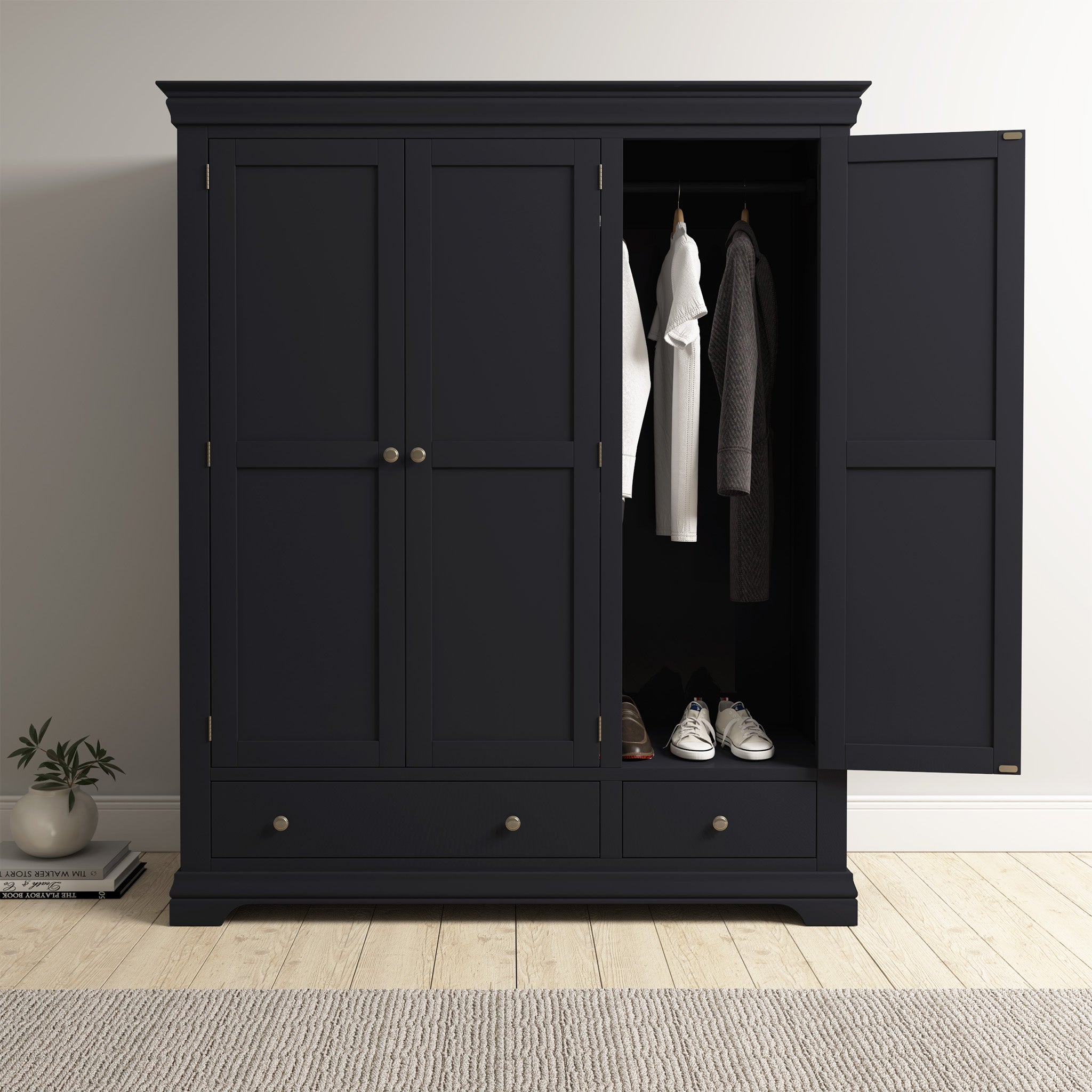 The Chalbury Triple Wardrobe in Dusky Black displays its roomy interior filled with clothes and shoes. Its modern design pairs well with the nearby plant and light wood floor, adding a stylish flair to any room.