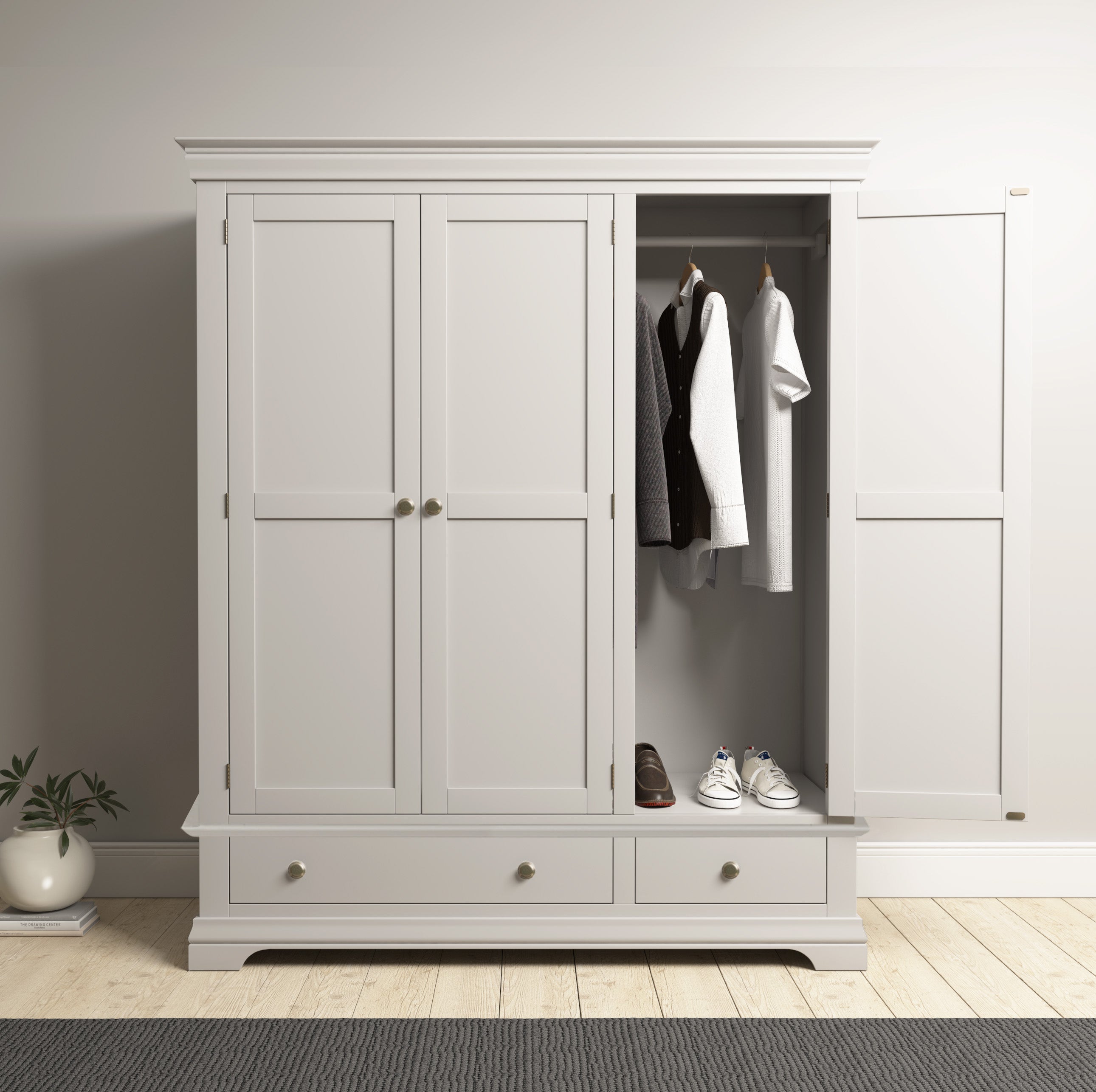 The Chalbury Triple Wardrobe in Pebble Grey stands open on a light wooden floor, showcasing neatly organized hanging clothes and shoes, with a plant nearby.