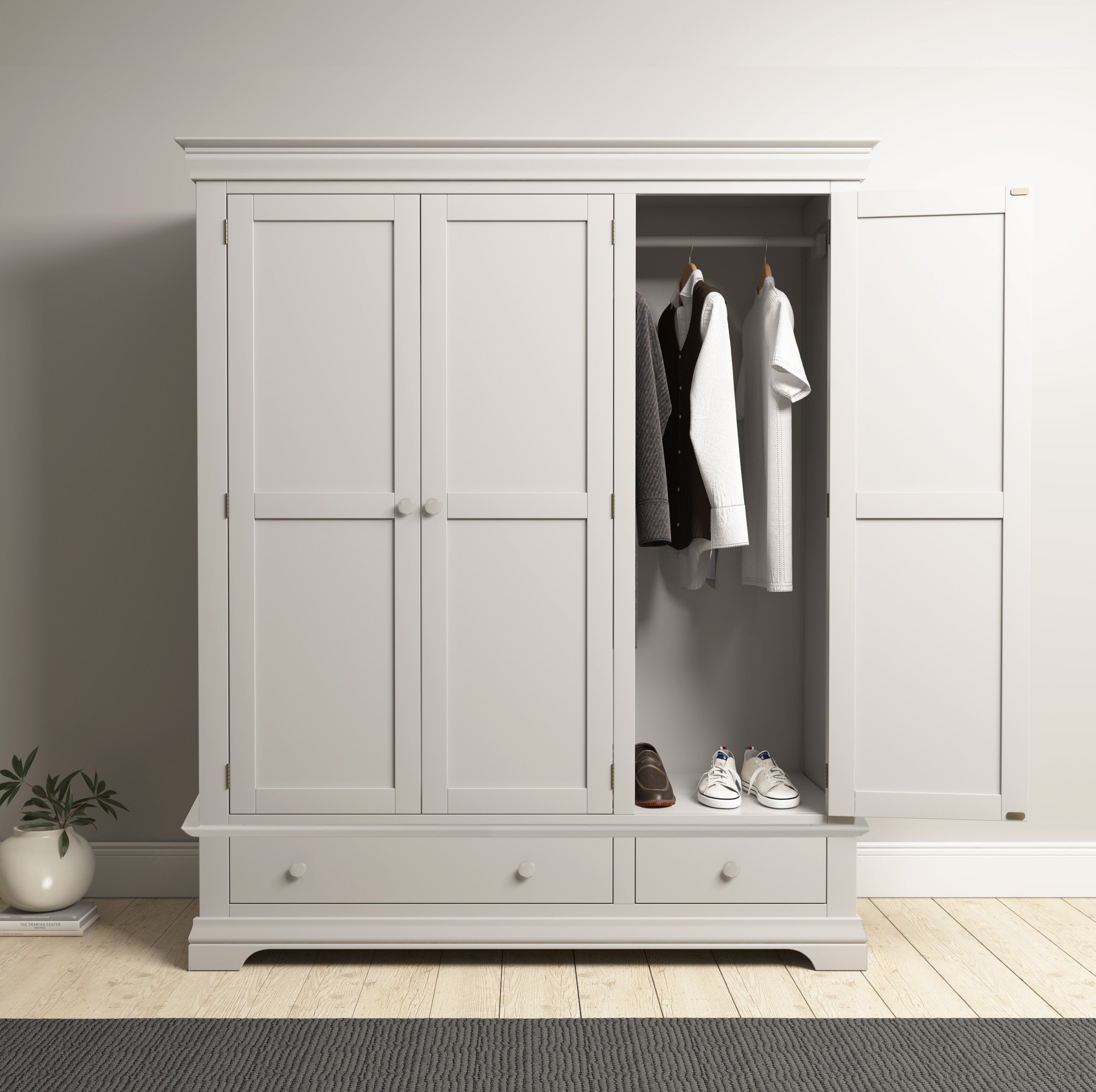 The Chalbury Triple Wardrobe in Pebble Grey, an ideal bedroom storage solution, has one door slightly open to reveal organized clothes, shoes, and two roomy drawers. A nearby plant adds a natural touch.