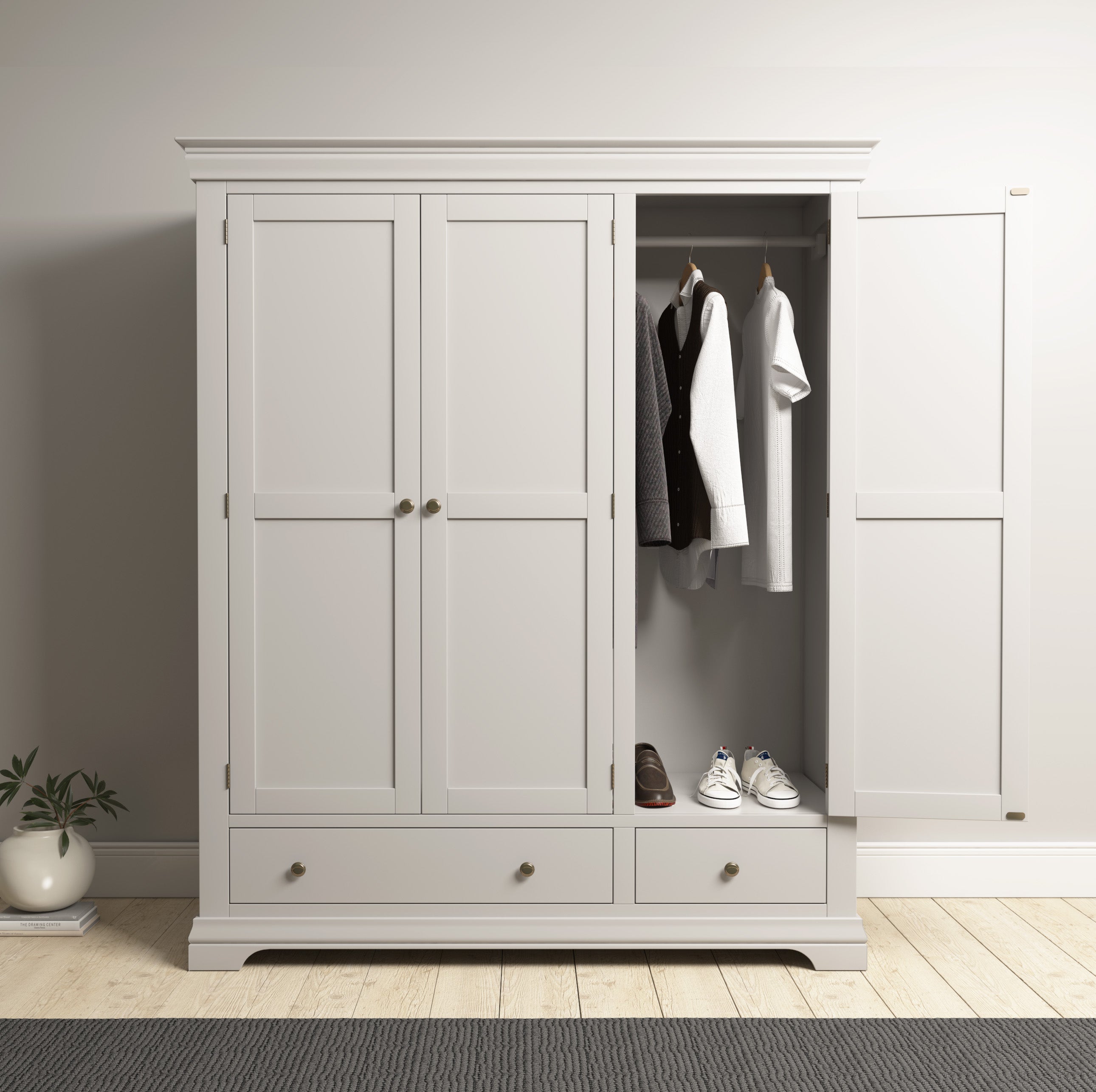 The Chalbury Triple Wardrobe in Pebble Grey enhances the bedroom with its doors open, showcasing neatly arranged clothes, shoes, and drawers. A small plant beside it adds a touch of nature to the serene setting.