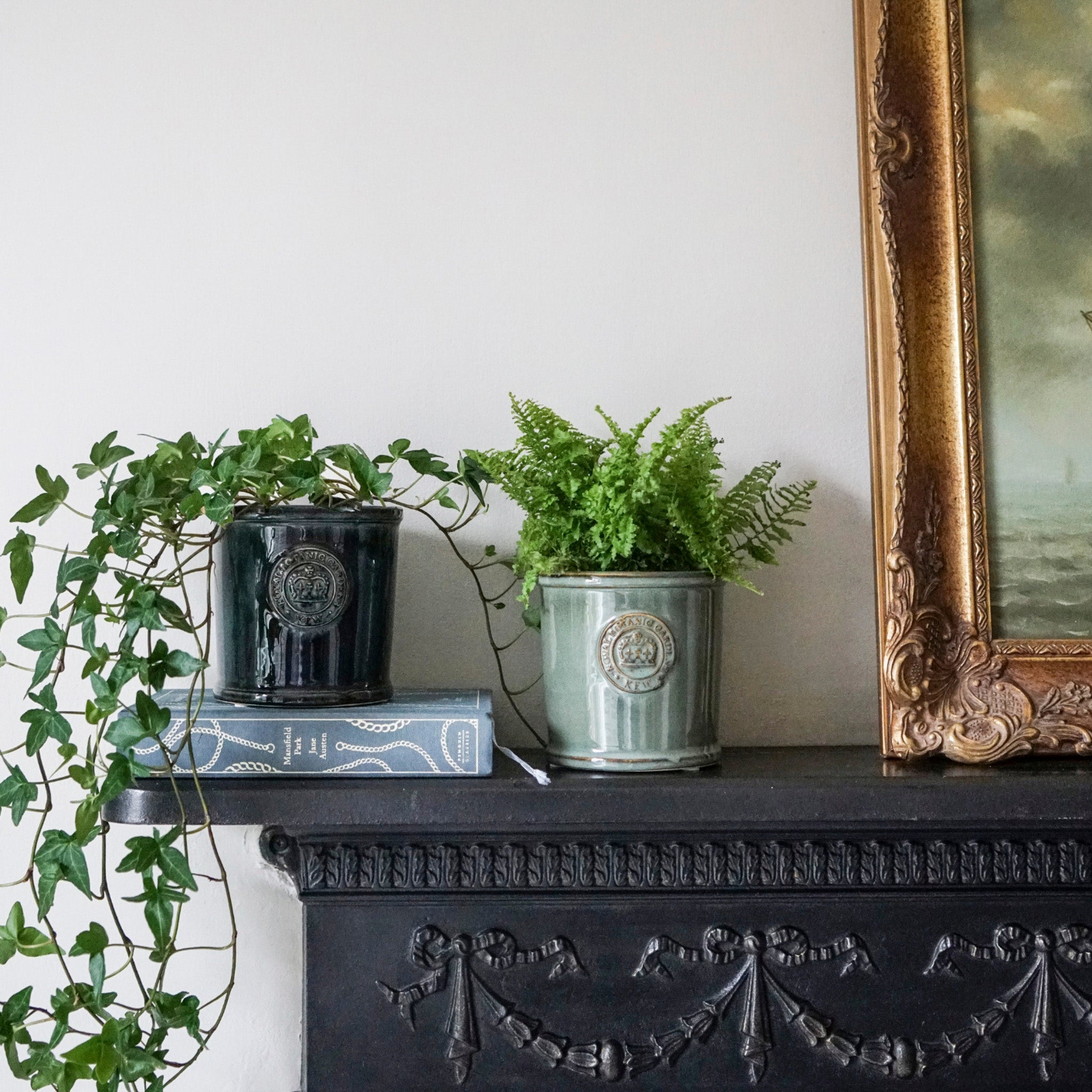 Two potted plants in Kew Reactive Glaze Sage Planters adorn the decorated mantel; one with trailing vines, the other a lush fern, set elegantly beside a framed painting.