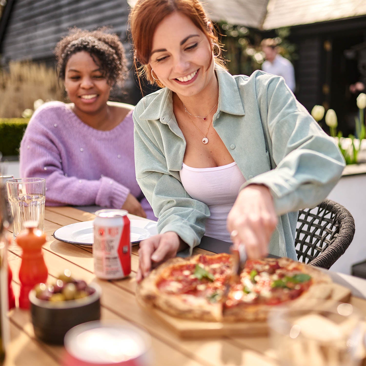 Two people enjoying an outdoor meal at the Kalama 8 Seat Rectangular Extending Dining Set with Teak Table in Charcoal; one slices a pizza while the other smiles. Drinks and food are spread on the table, complete with stylish woven chairs.