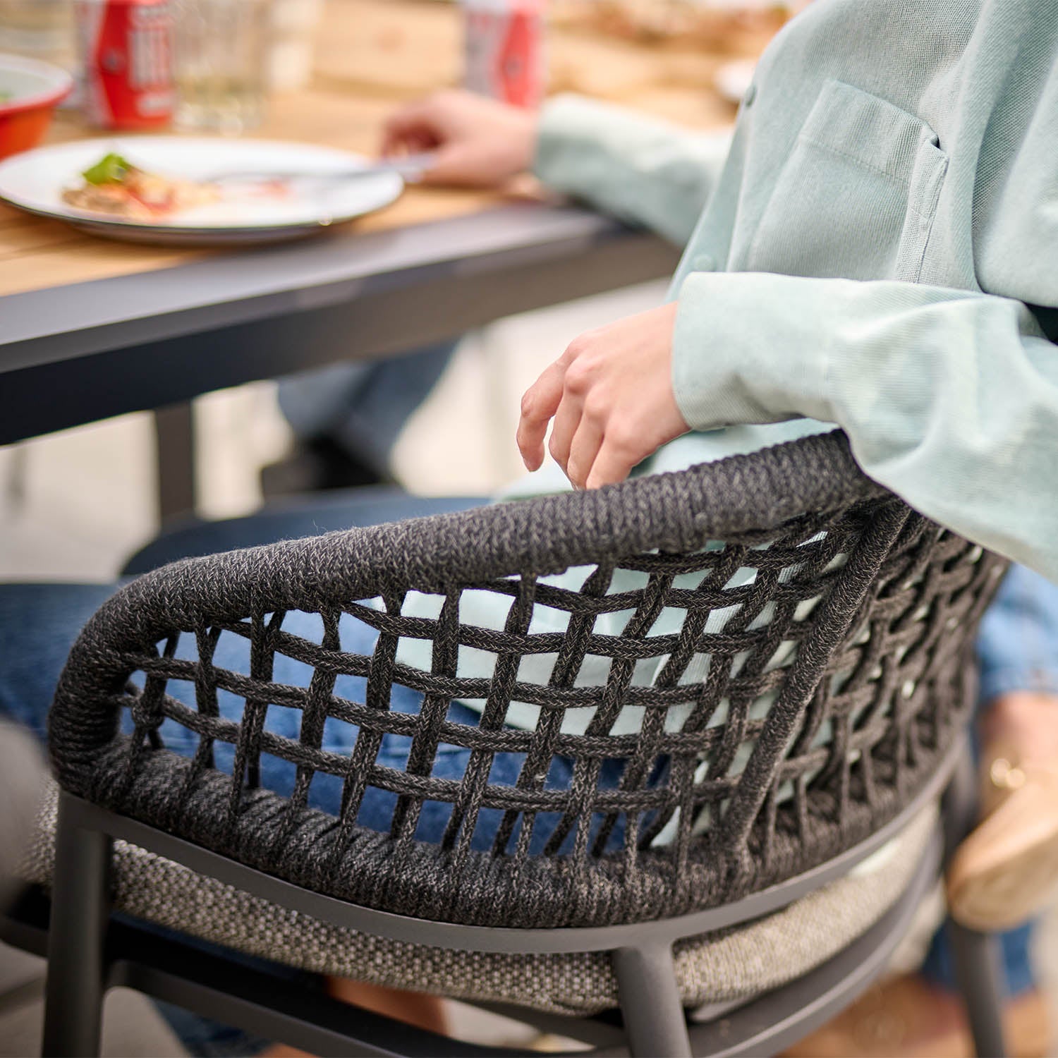 Casual dining scene with a person in a green shirt sitting on one of the woven chairs from the Kalama 8 Seat Rectangular Extending Dining Set with Teak Table in Charcoal, beside a teak dining table adorned with plates and beverages.