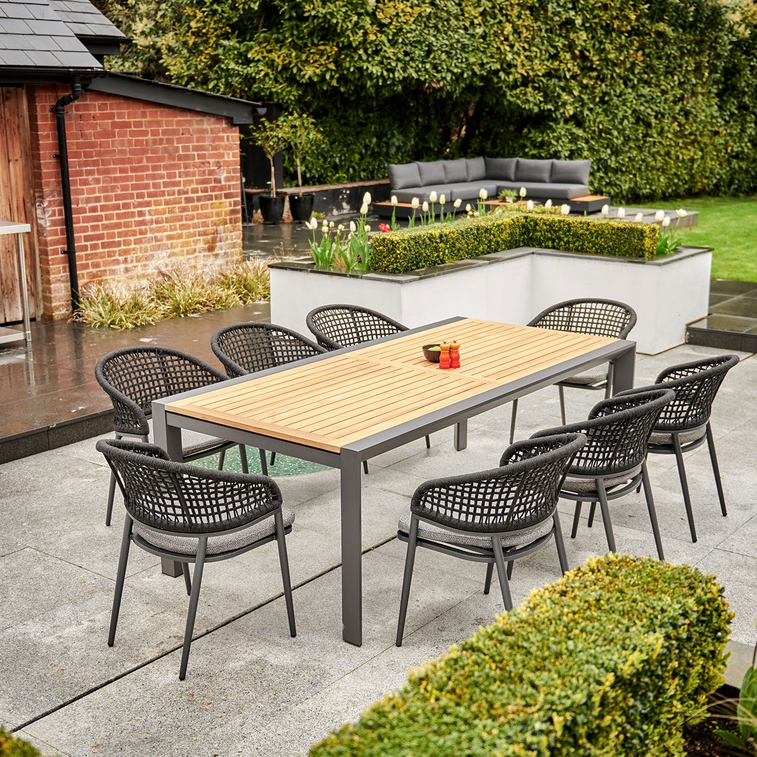 Outdoor patio with a Kalama 8 Seat Rectangular Extending Dining Set featuring a teak table in charcoal surrounded by woven chairs, a brick wall, and greenery in the background.