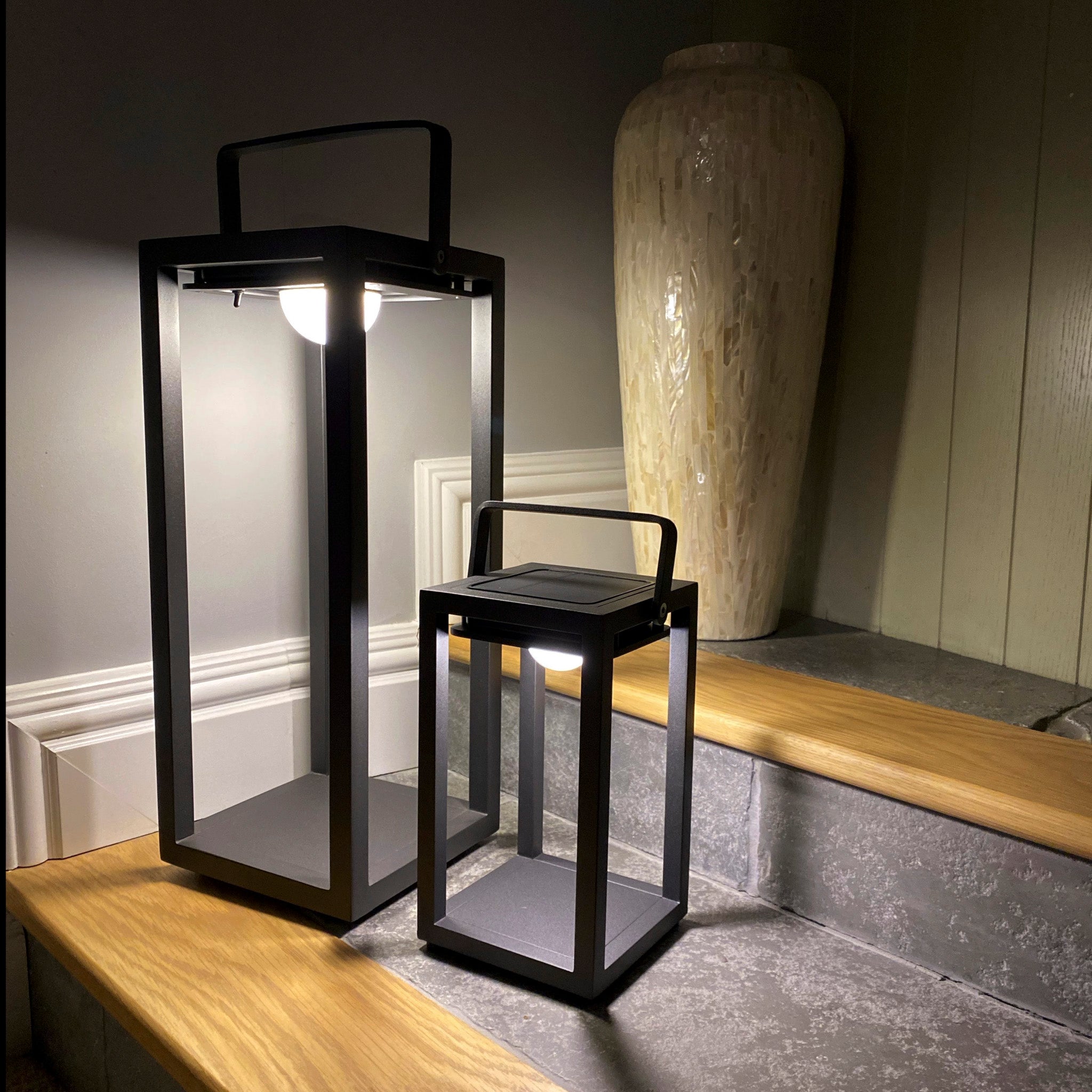 Two Luxor Small Table Outdoor and Indoor Solar Lanterns in Charcoal, one large and one small, grace wooden steps beside a large decorative vase. These weatherproof lights are designed to withstand the elements while adding charm to any setting.